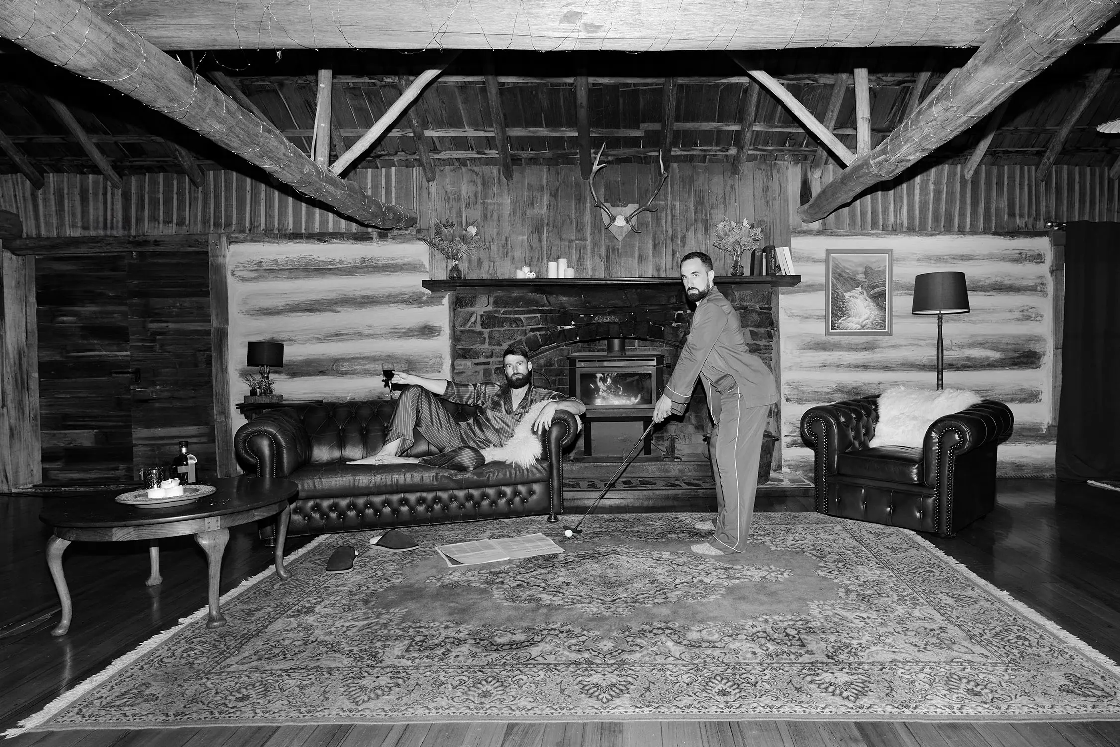 Inside a richly decorated wood log cabin, with a burning fireplace, leather couches and a rich rug. One man lounges on a sofa with a glass of wine while the other plays minigolf - both in bare feet and silk pajamas.