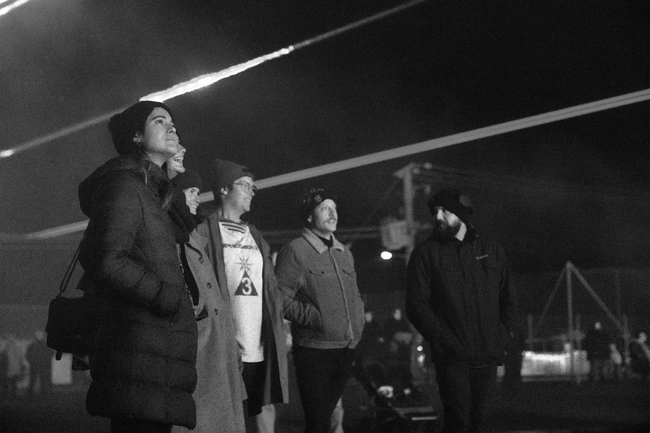 A group of five people, wearing winter clothing, are looking up at the night sky, illuminated by streaks of light, surrounded by a misty atmosphere