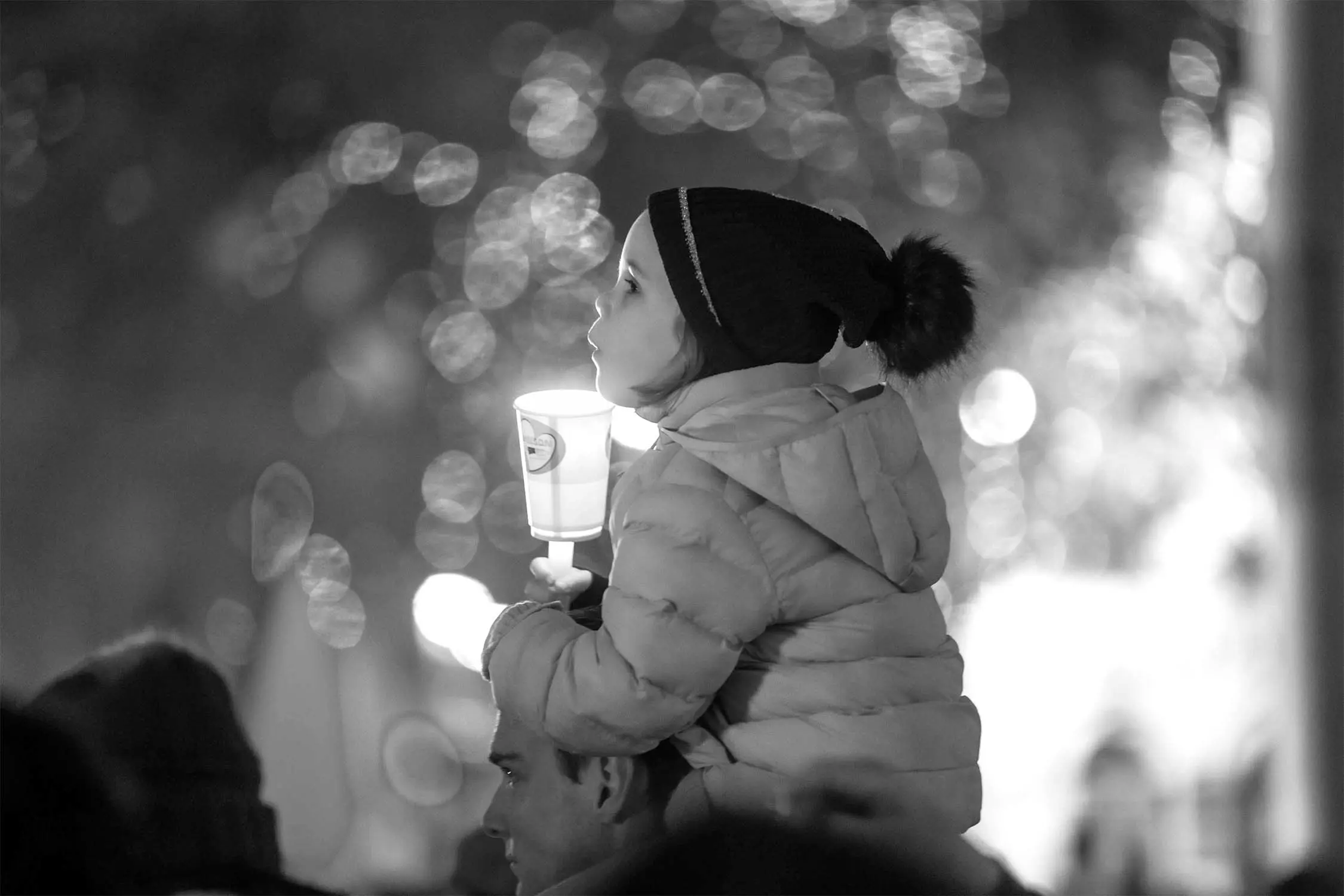 A child in winter clothing sits on an adult's shoulders, holding a lit cup and gazing up, with blurred lights creating a festive black-and-white background.