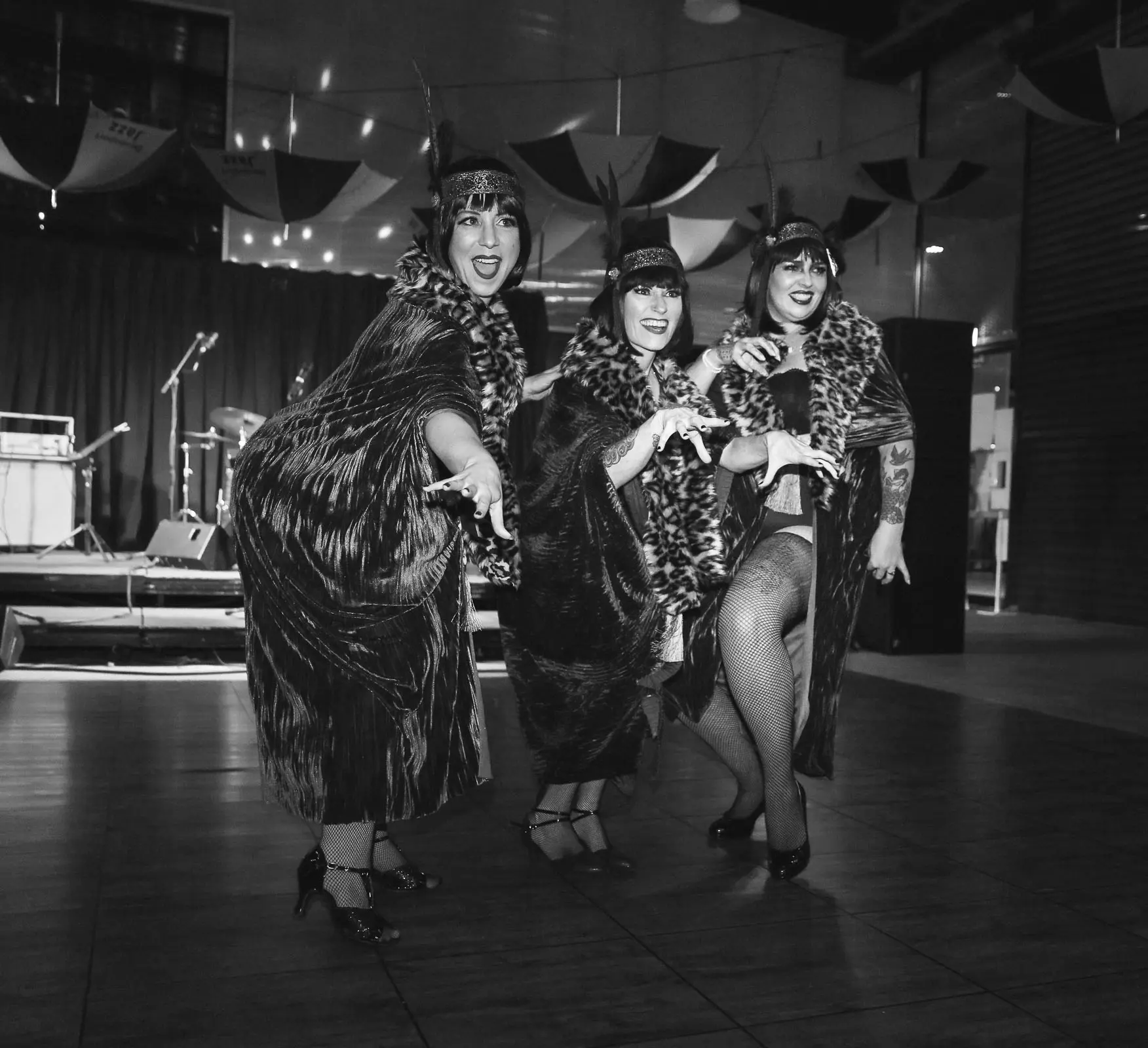 Three burlesque performers, wearing slinky robes and leopard print furs, sing and dance on an open dancefloor stage.