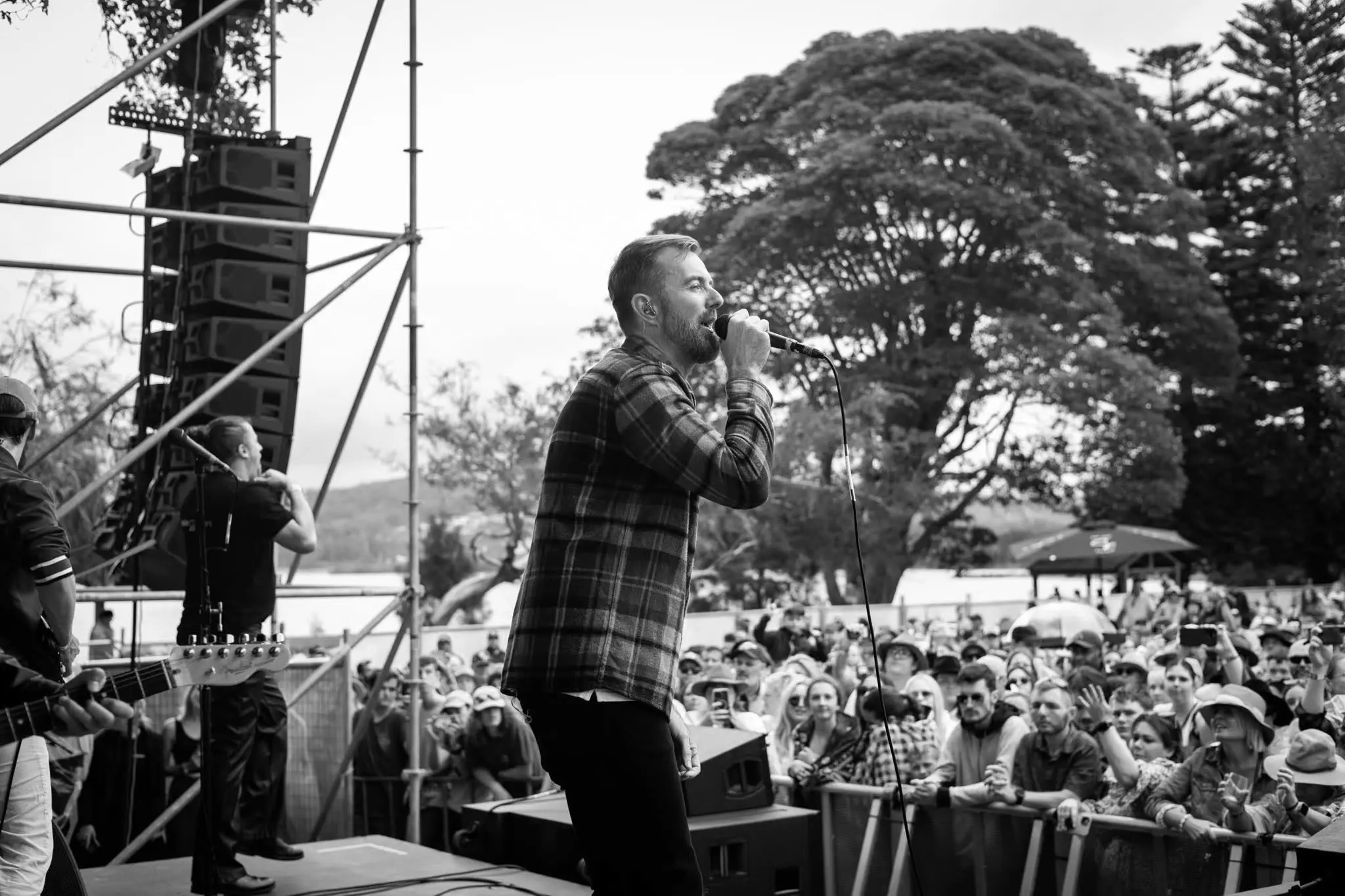 A band performs on an outdoor festival stage, featuring the lead singer at the front, looking out to the crowds of people in front.