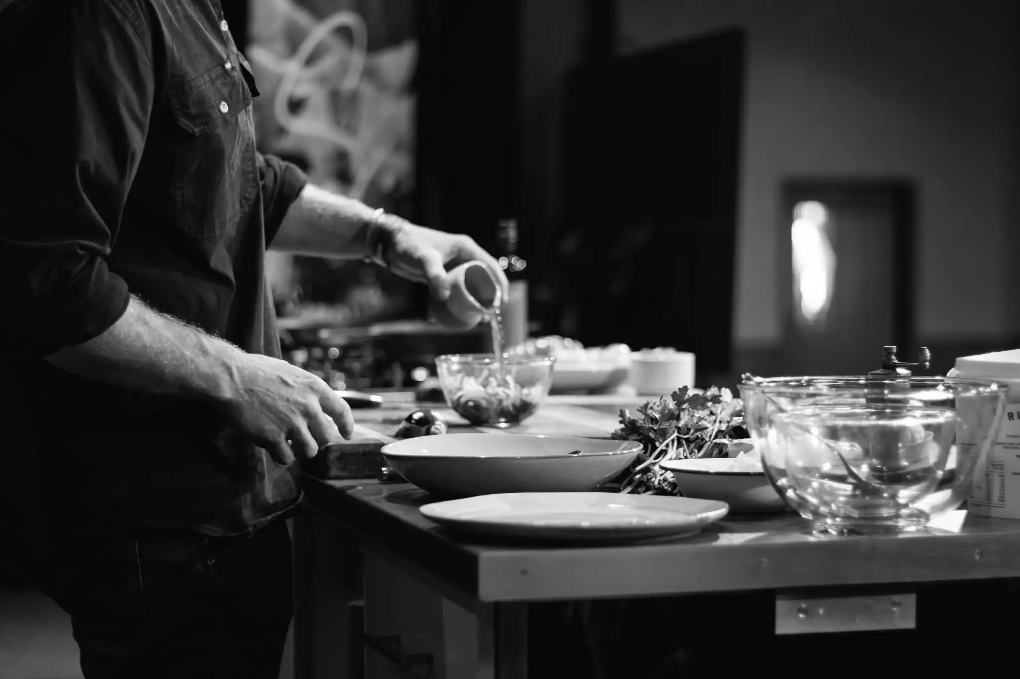 A man stands at a benchtop table, demonstrating the preparation of food. The bench is laden with plates, condiments, herbs and other cooking implements.