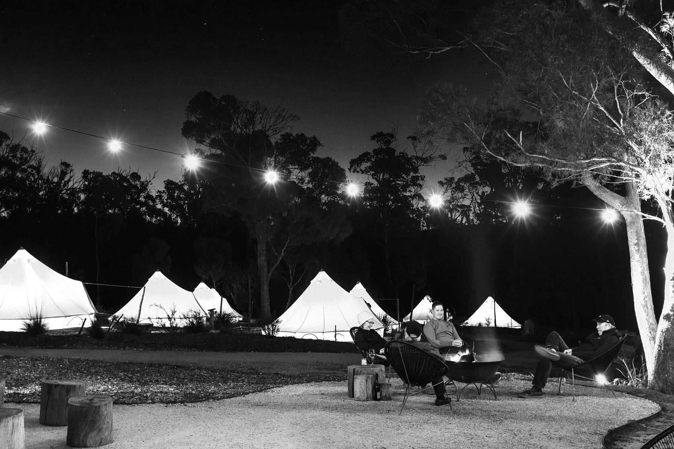 Several canvas tents lit up from the inside are dotted amidst the bush at dusk, with people sitting around a campfire under fairy lights strung between trees.