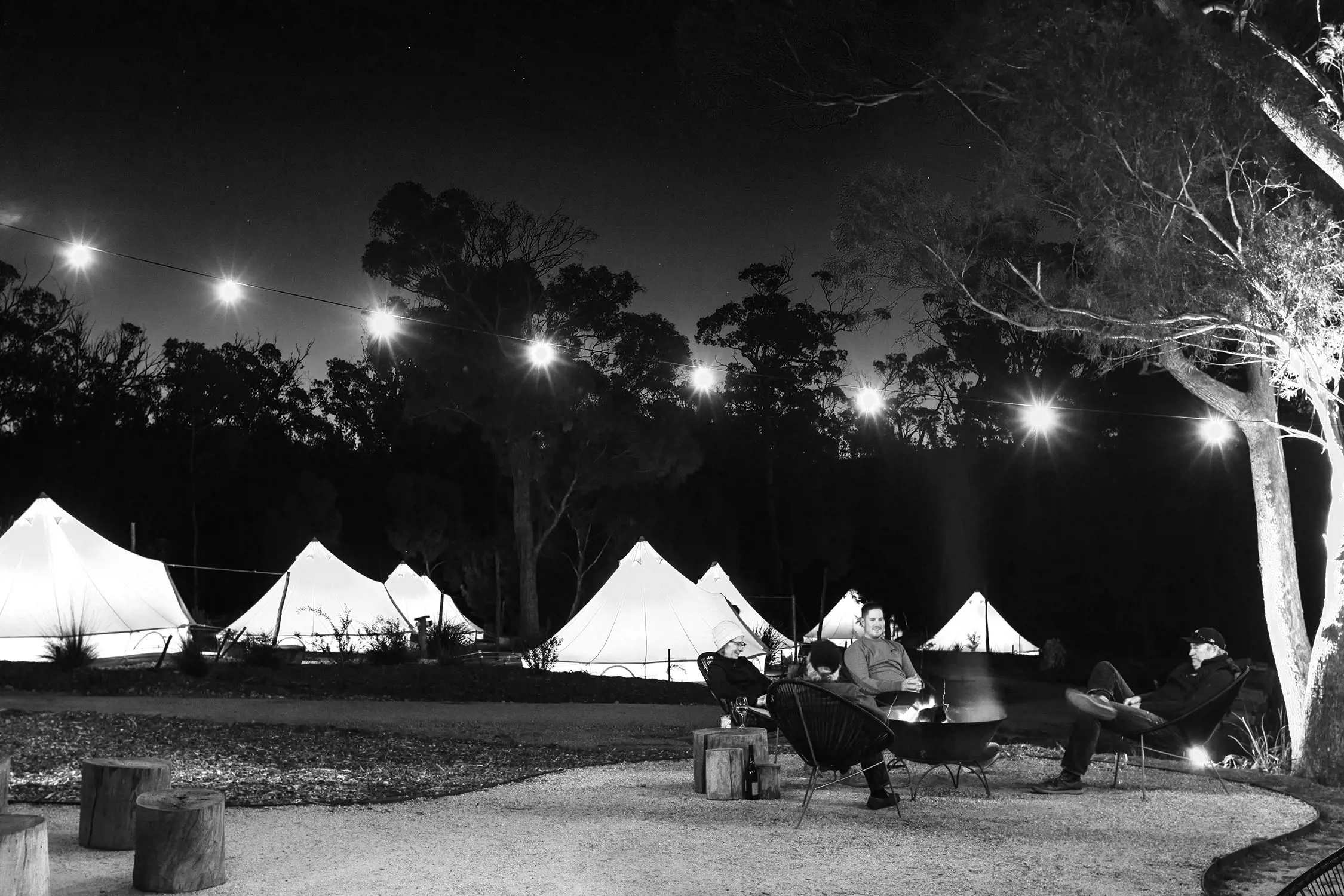 Several canvas tents lit up from the inside are dotted amidst the bush at dusk, with people sitting around a campfire under fairy lights strung between trees.