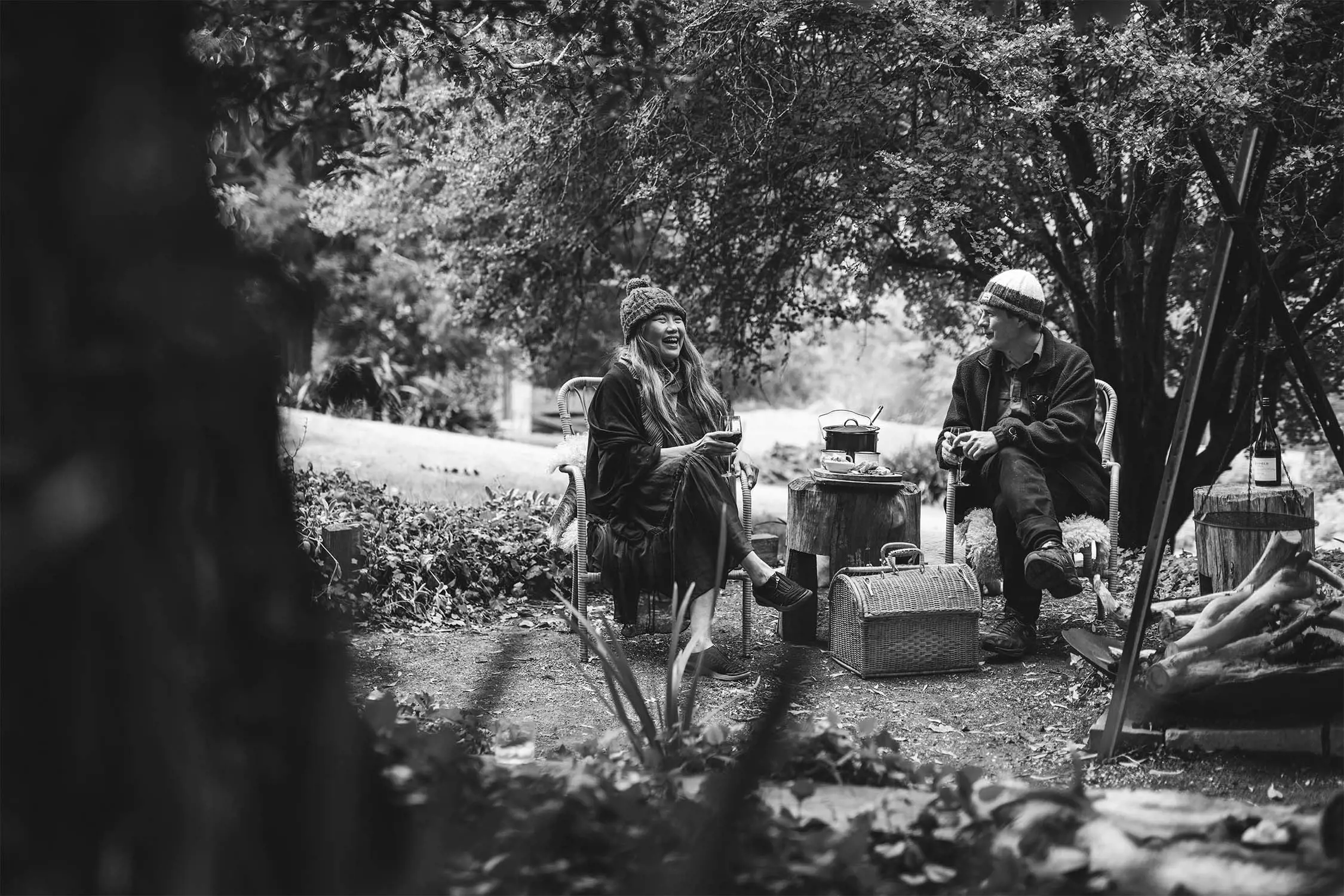 Two people sitting outside, one laughing while holding a cup, surrounded by greenery and a picnic setup, captured in black and white.