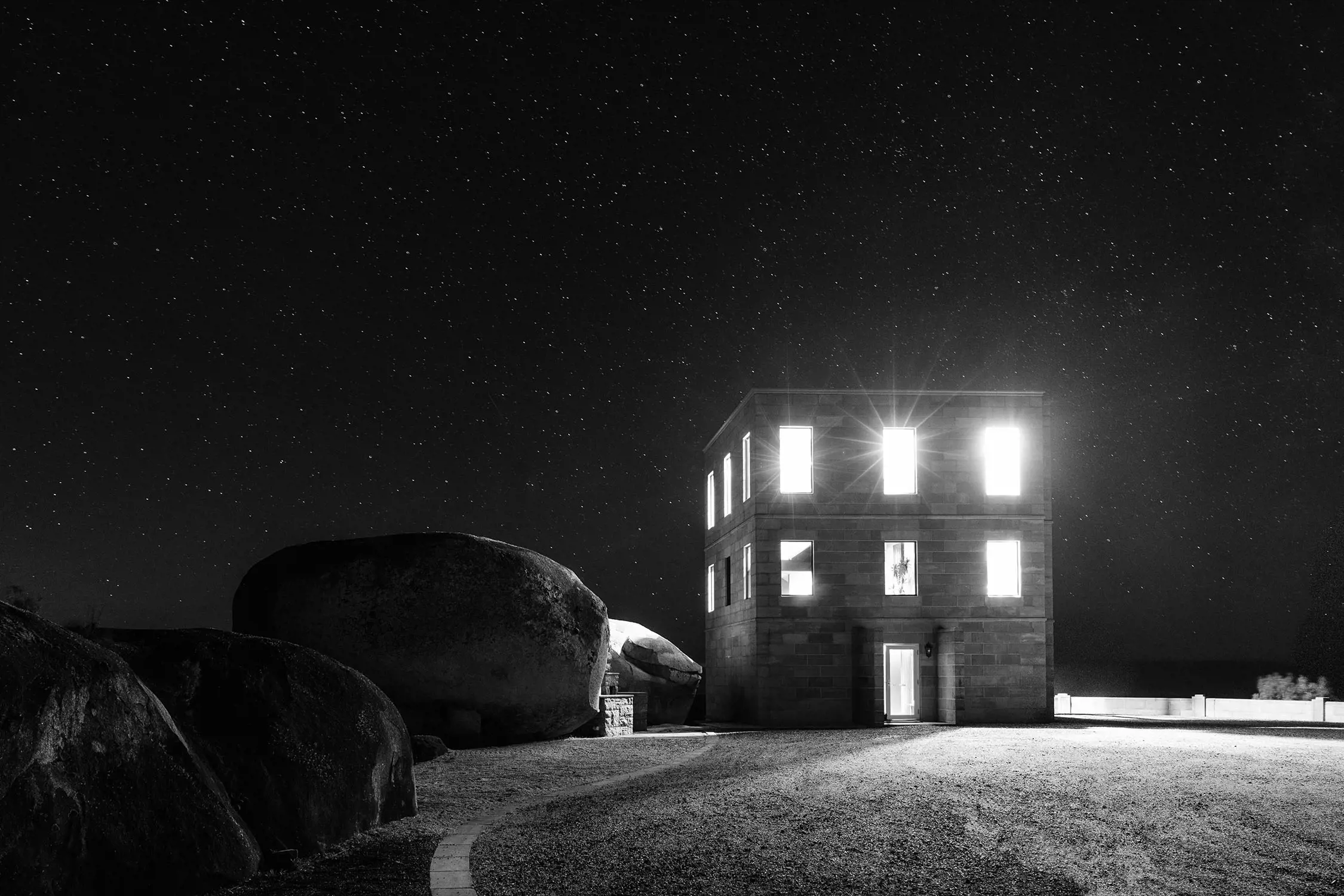 Against a dark black sky, bright light is blazing from the windows of a three-storey castle-like building perched among huge boulders.