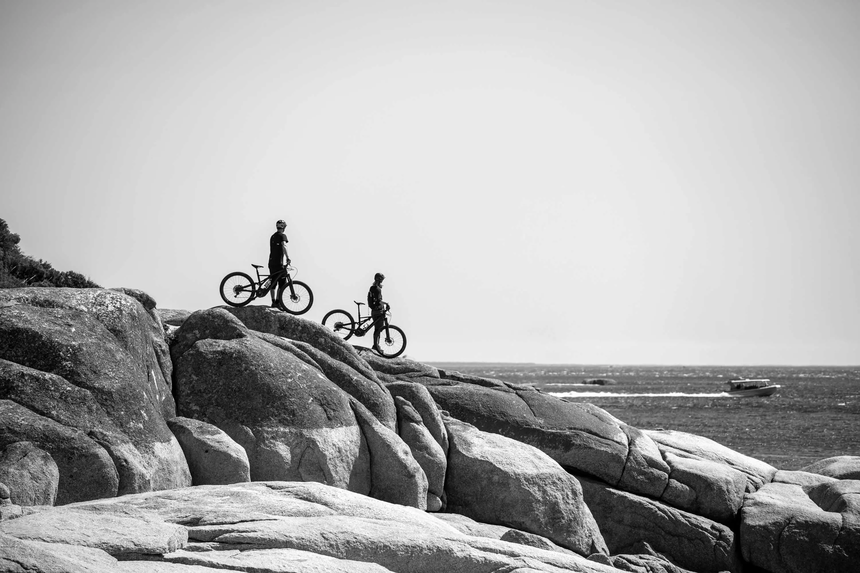 Two mountain bike riders stop on the top of a large, flat rock at the shore, looking out to the ocean.