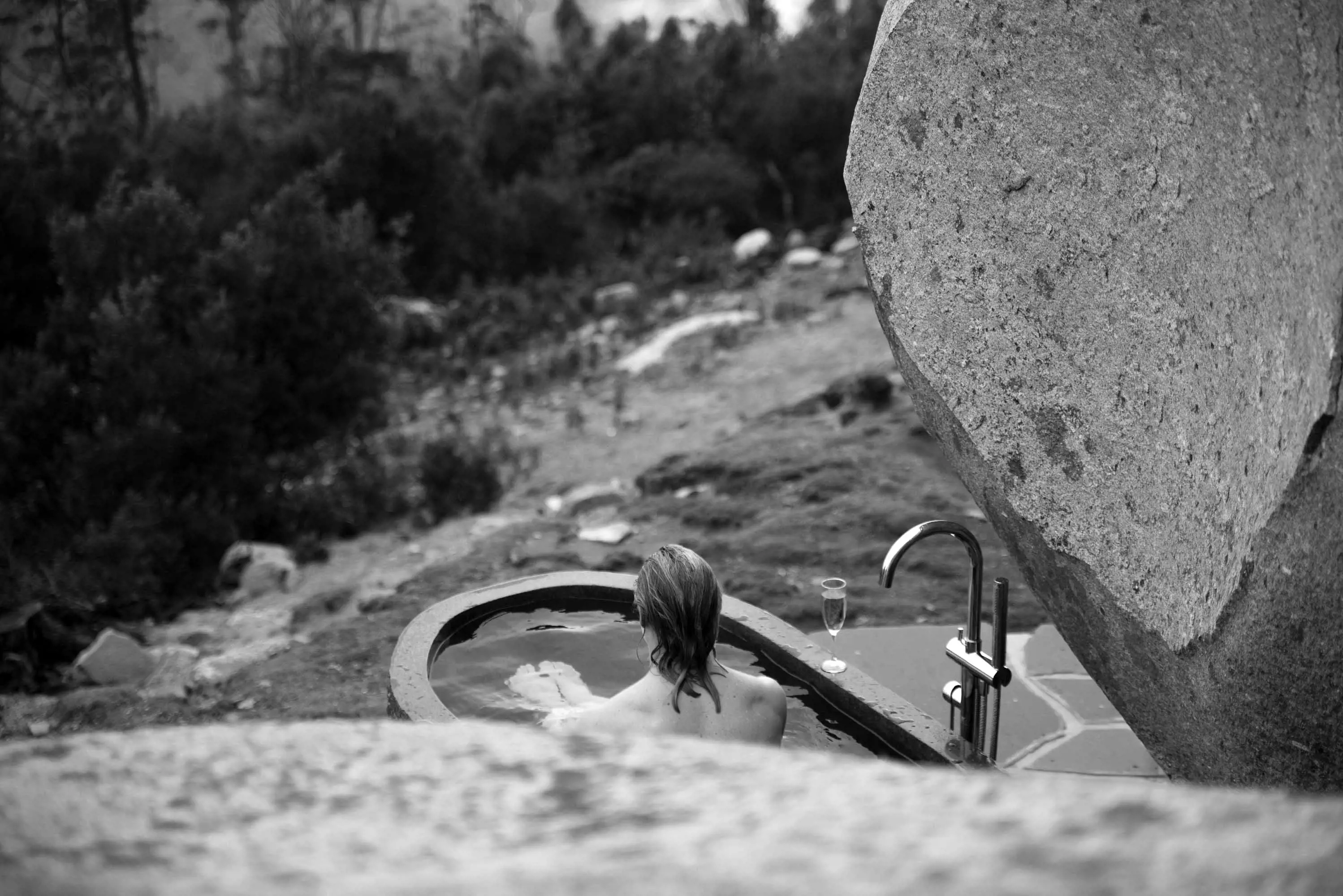 A woman relaxes in a large, stone bath in the shade of large rocks, overlooking bushland.