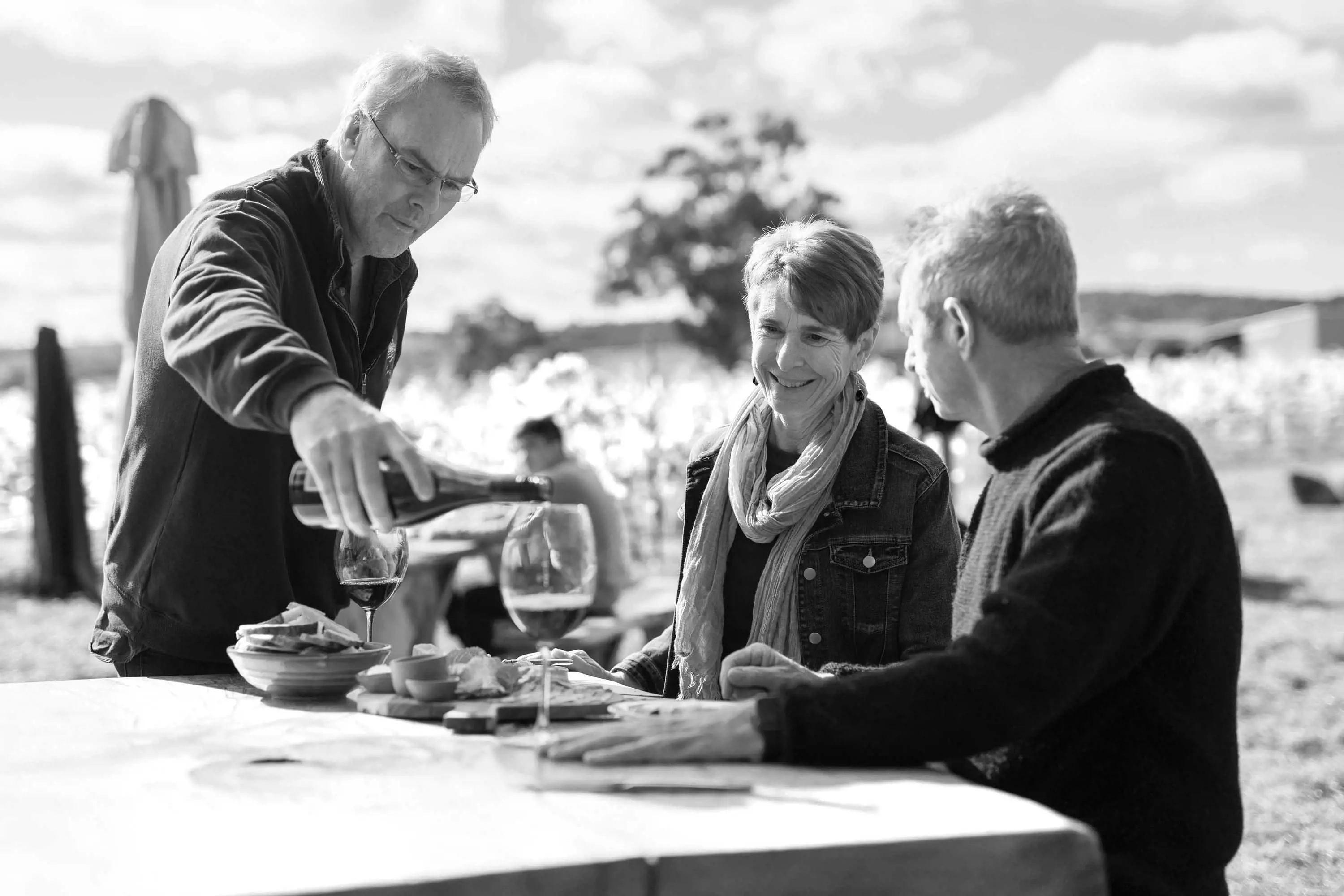 A retired couple sit at a wooden table outside and are served wine by a winemaker.