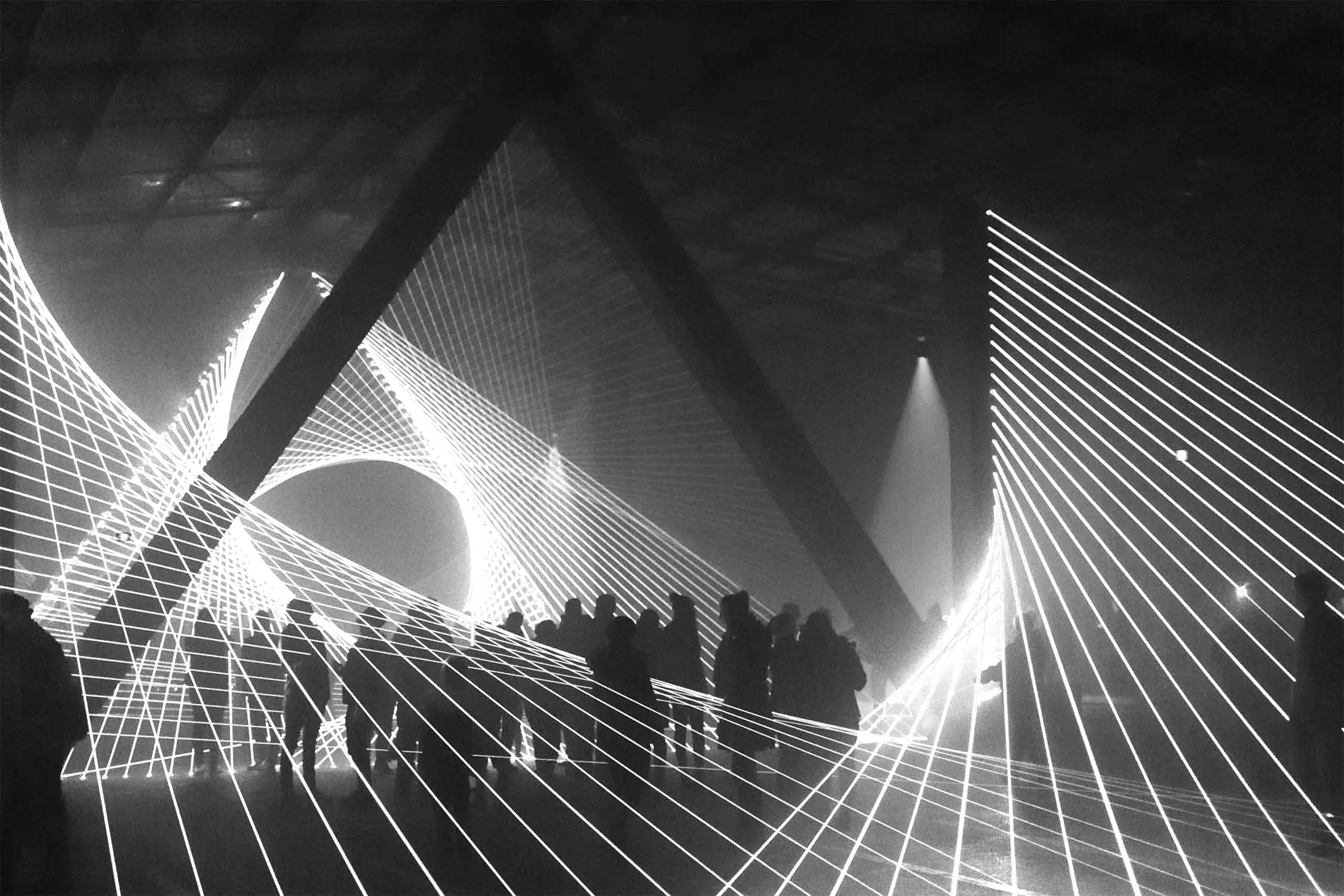 Silhouettes of people standing beneath geometric light patterns in a darkened space, creating a dynamic visual effect, captured in black and white.
