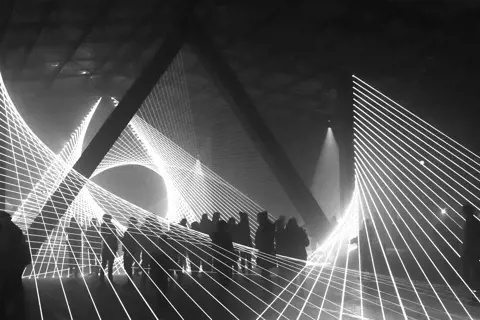 Silhouettes of people standing beneath geometric light patterns in a darkened space, creating a dynamic visual effect, captured in black and white.