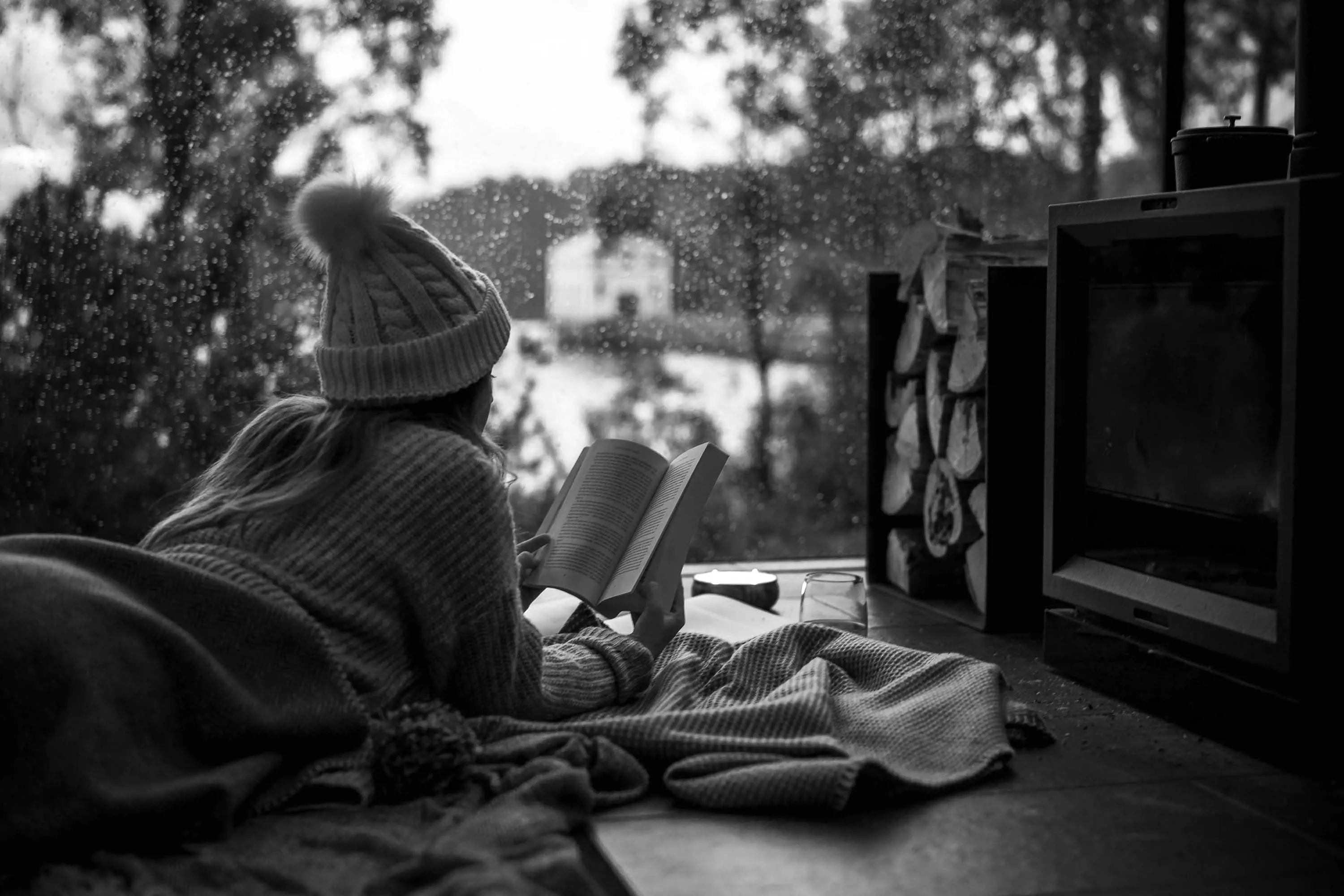 A young woman wearing a beanie and wrapped in a blanket reads a book lying on a rug beside a fire, with a lake seen through a large window in in the background.