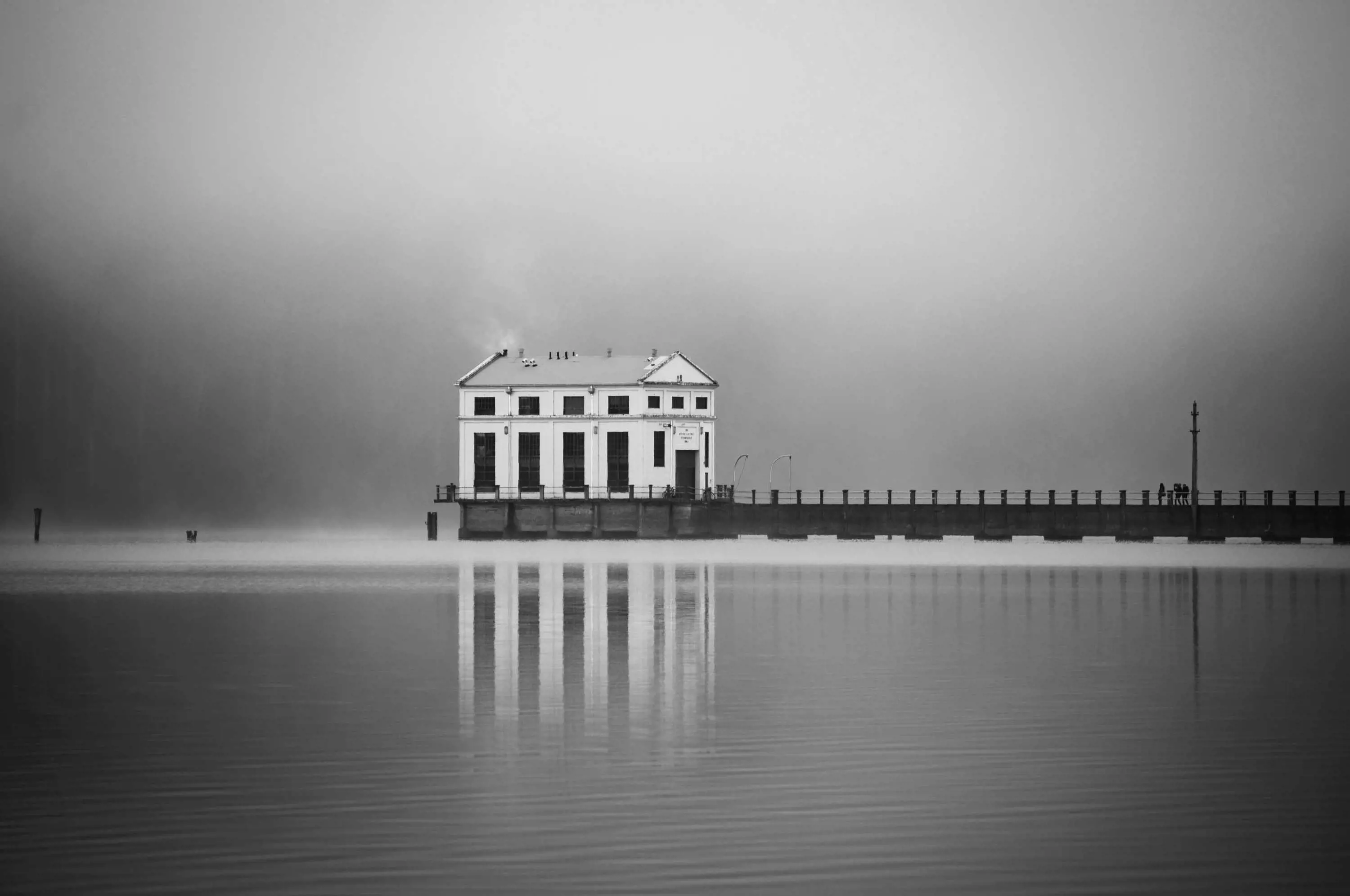 An old, white, shed like building at the end of a long pier is reflected in the still water of a lake.
