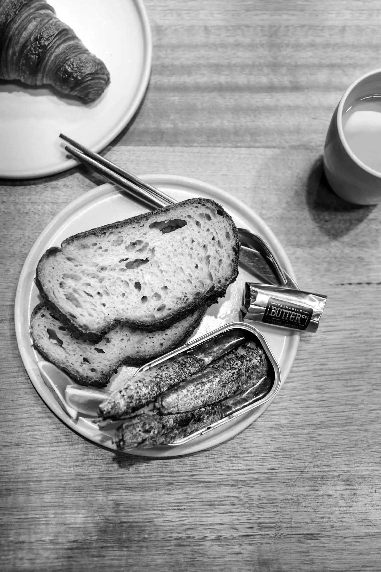 Sliced bread, sardines in a tin and btter sit on a white ceramic plate on a wooden table with other baked goods.