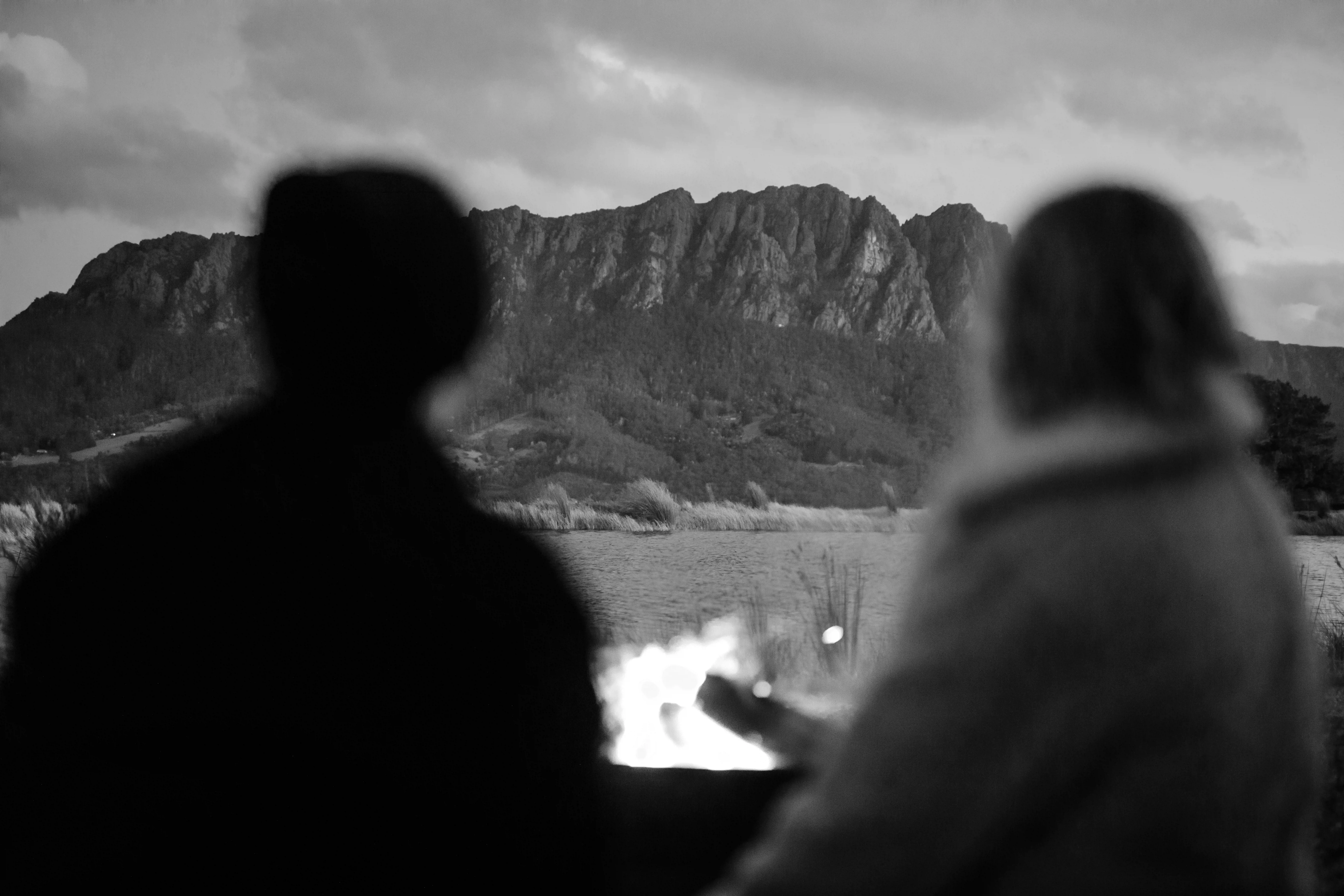 A couple sit in the foreground in front of a fire and gaze at a mountain range in the distance.