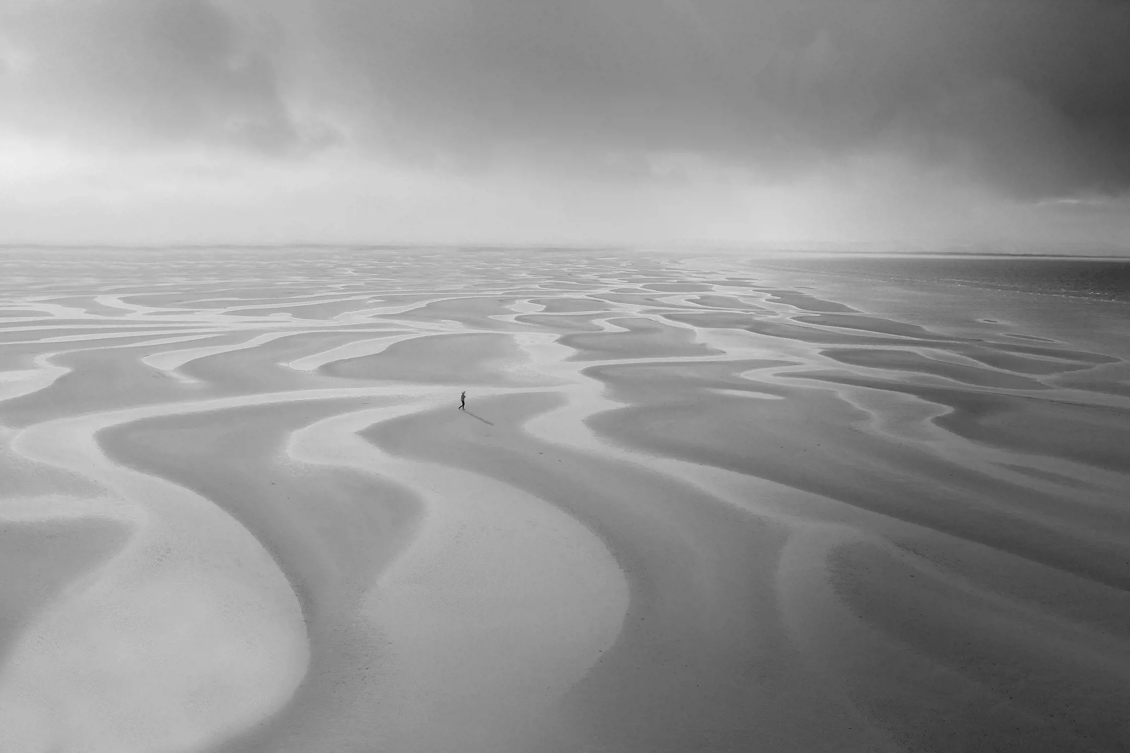 An otherworldly, desert-like sandy beach is covered in ripples made by the wind. A tiny figure stands on the beach and dark clouds loom overhead.