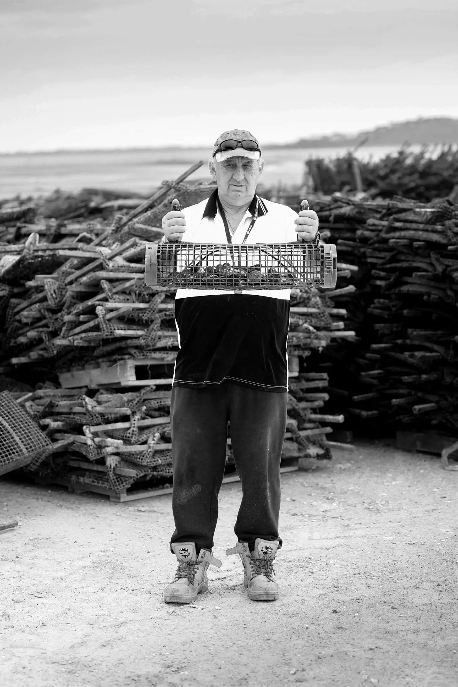 A man holding a fish trap in front of a stack of crates, with a landscape in the background, captured in black and white.