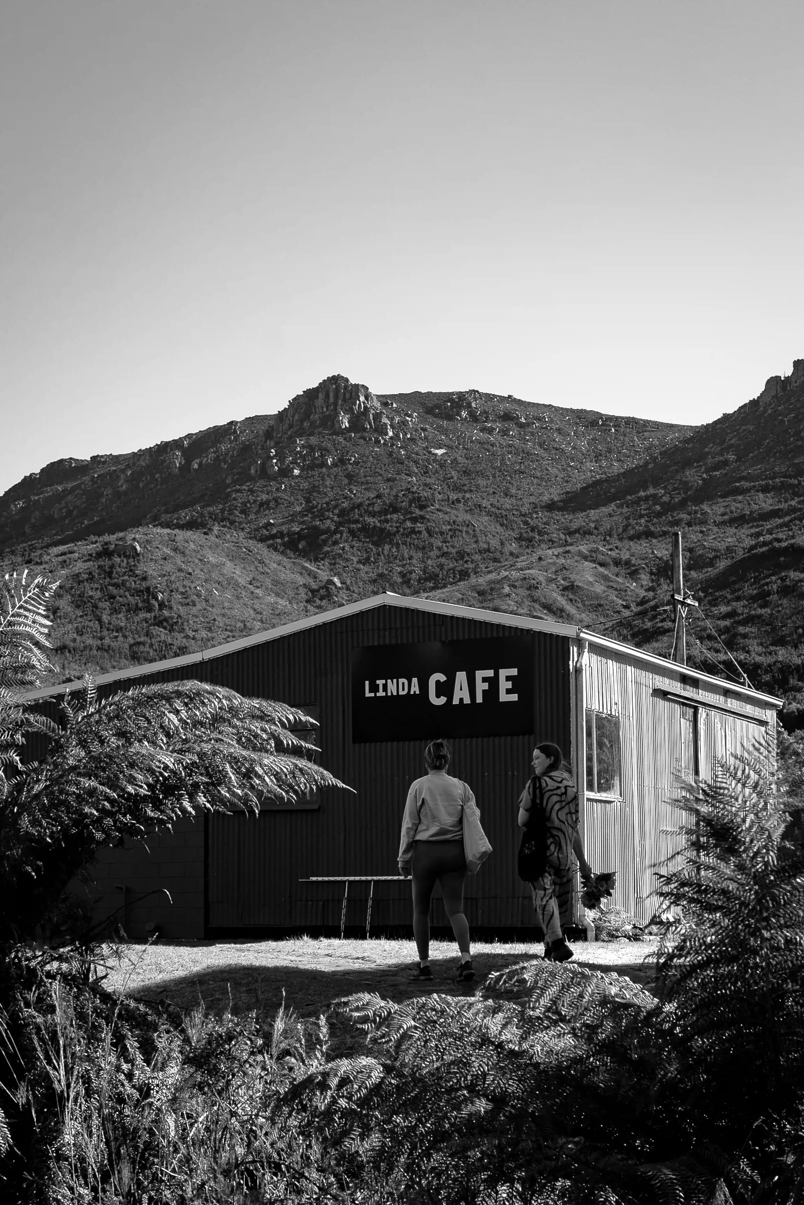 Two people stand on a wooden deck at the side of a large wooden shed-like structure with the words Linda Cafe hand-painted on one side.