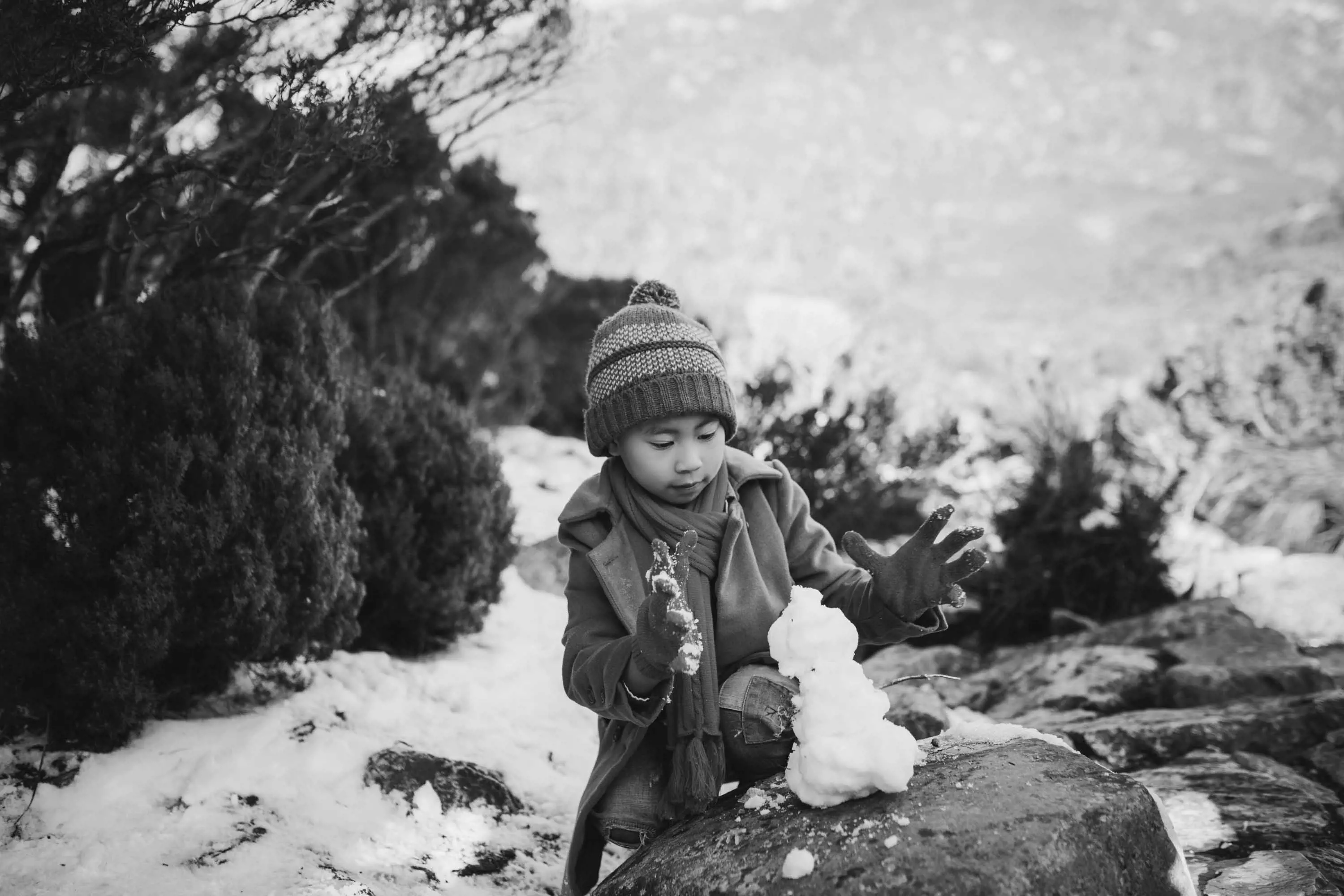 A young boy wearing a knitted beanie and a heavy winter coat makes a snow man on a large stone.