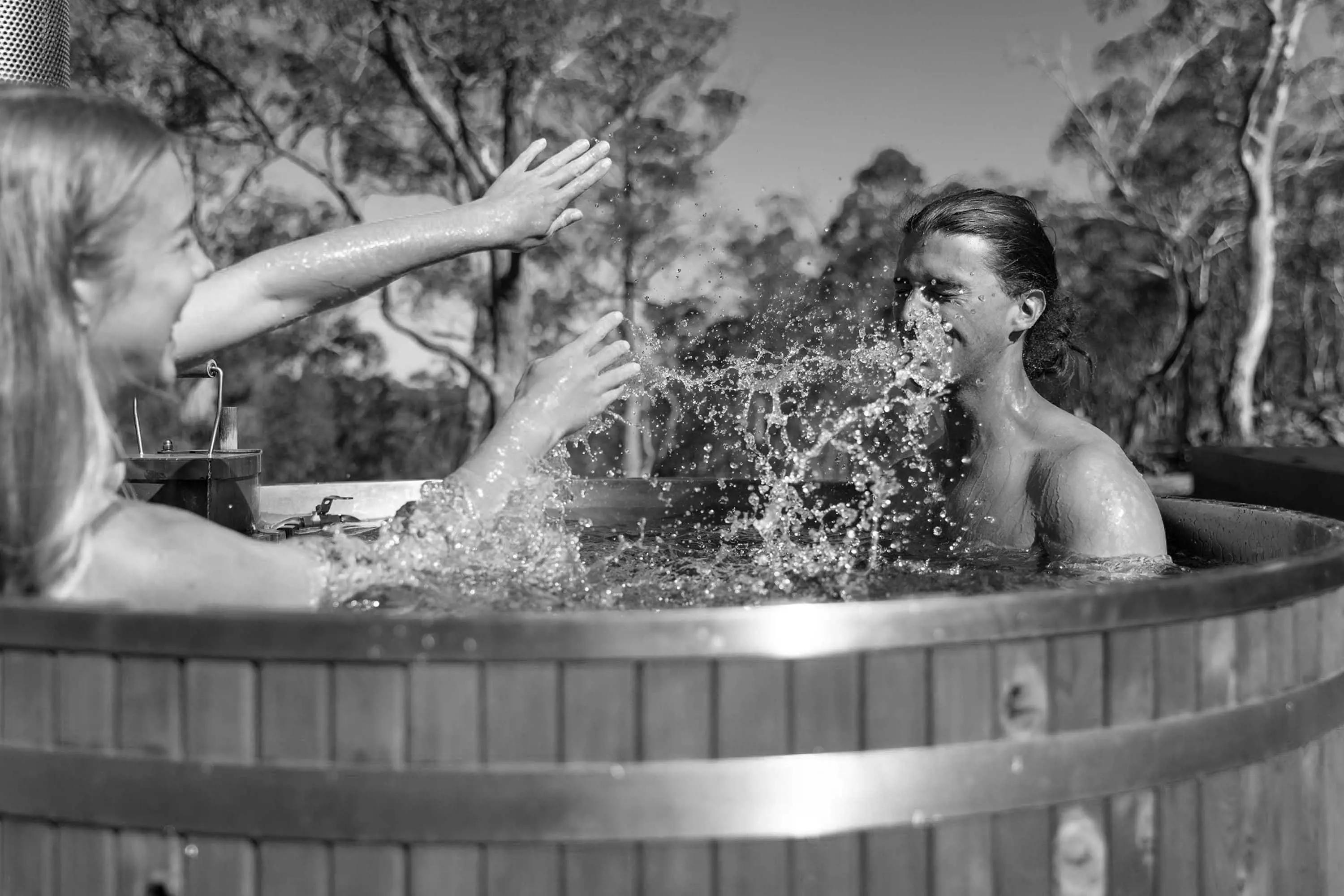 A young couple splash water in a large, outdoor hot-water tub made of wood.