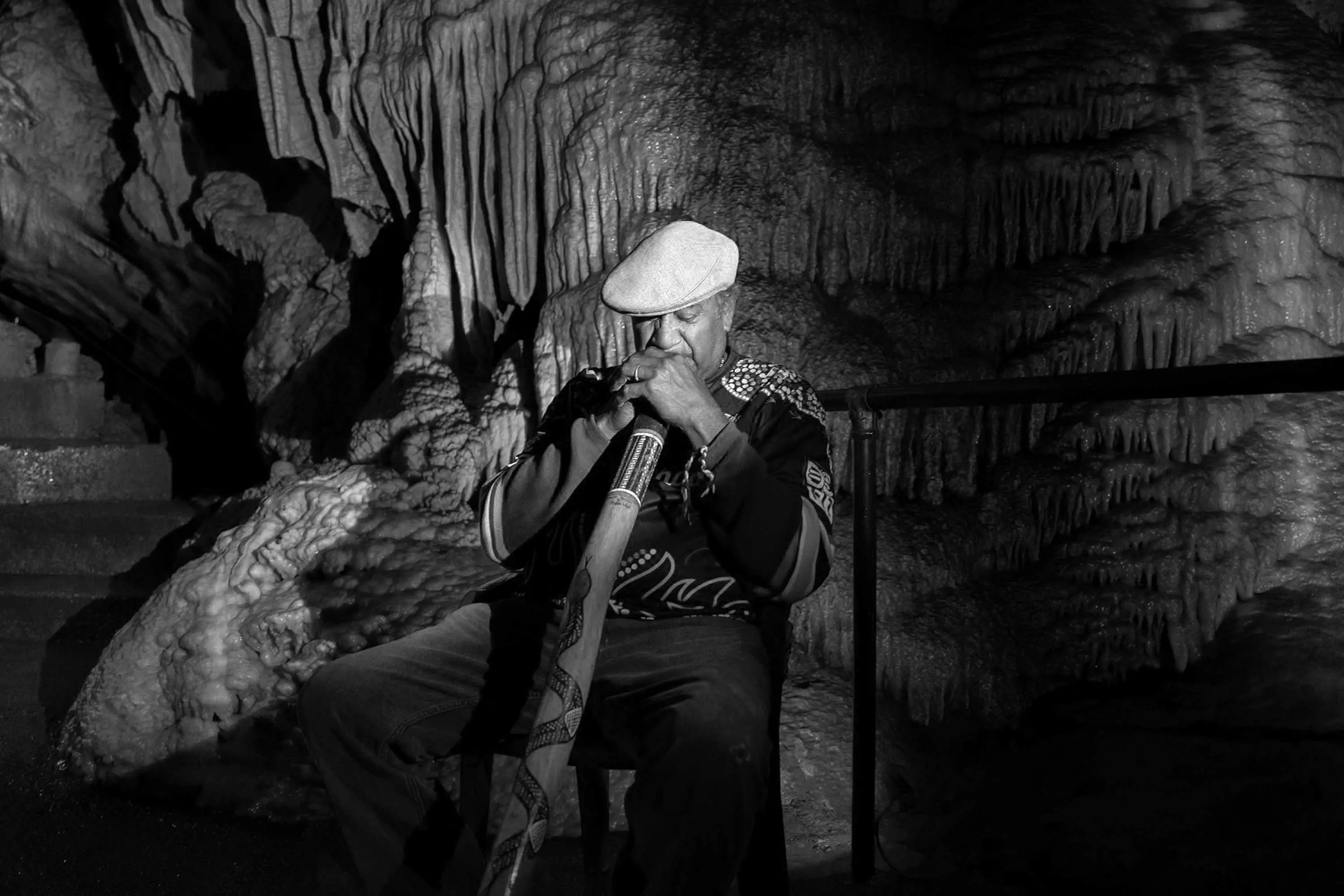 A man wearing a flat cap and a woolen jumper plays a wooden didgeridoo in limestone caves.