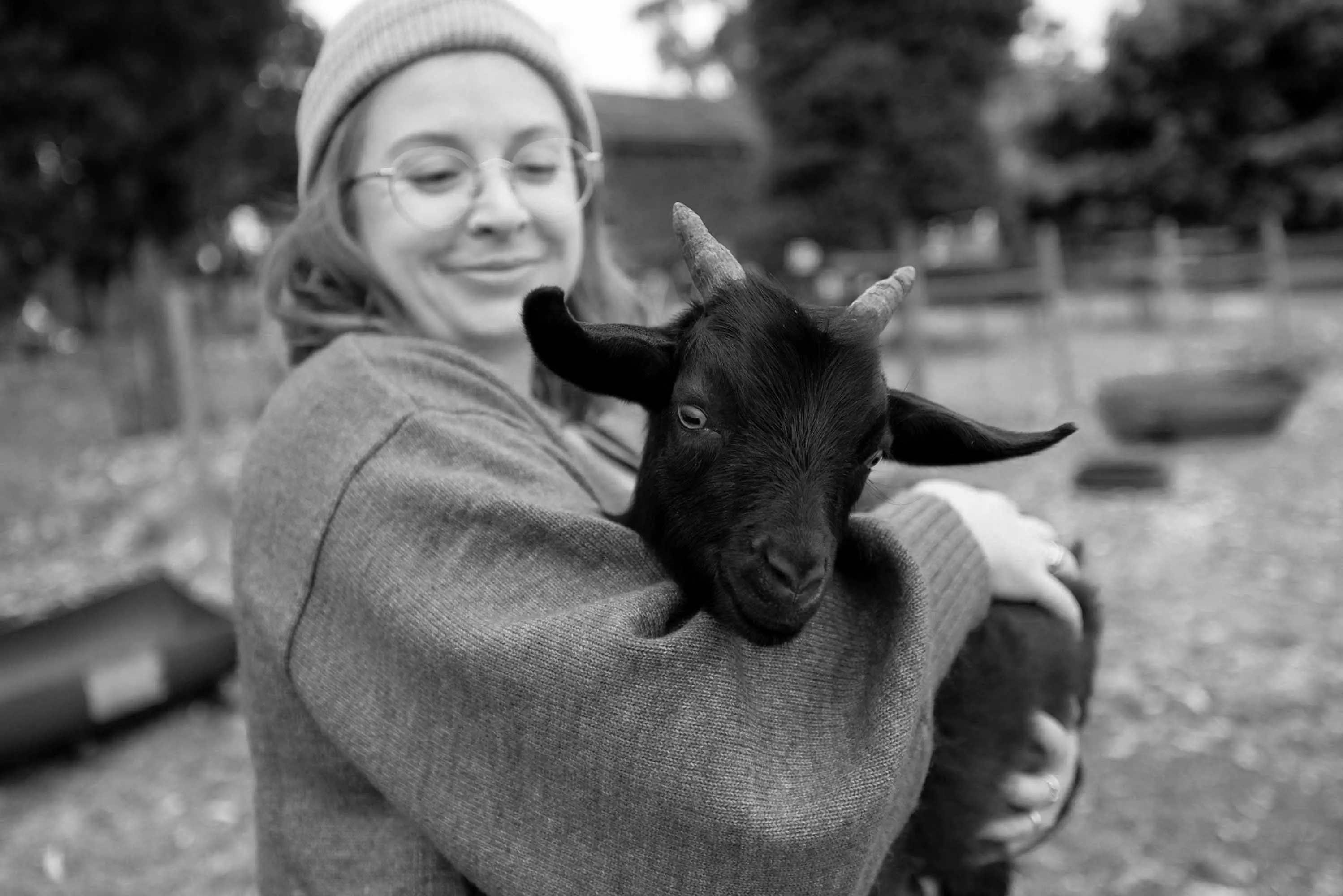 A woman wearing glasses and a knitted beanie holds a small, black goat.