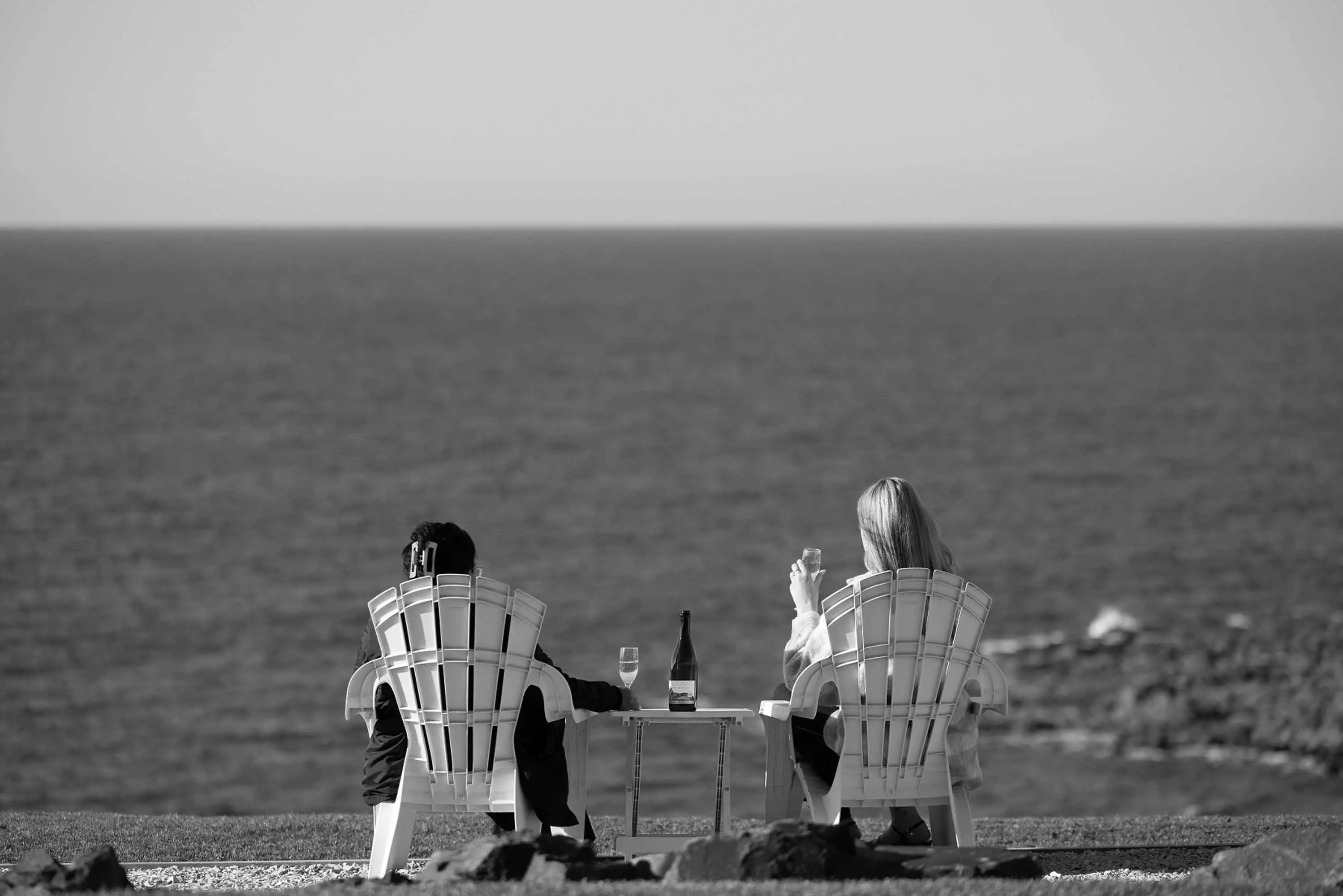 A couple sit in tall outdoor recliners next to the ocean and drink wine.