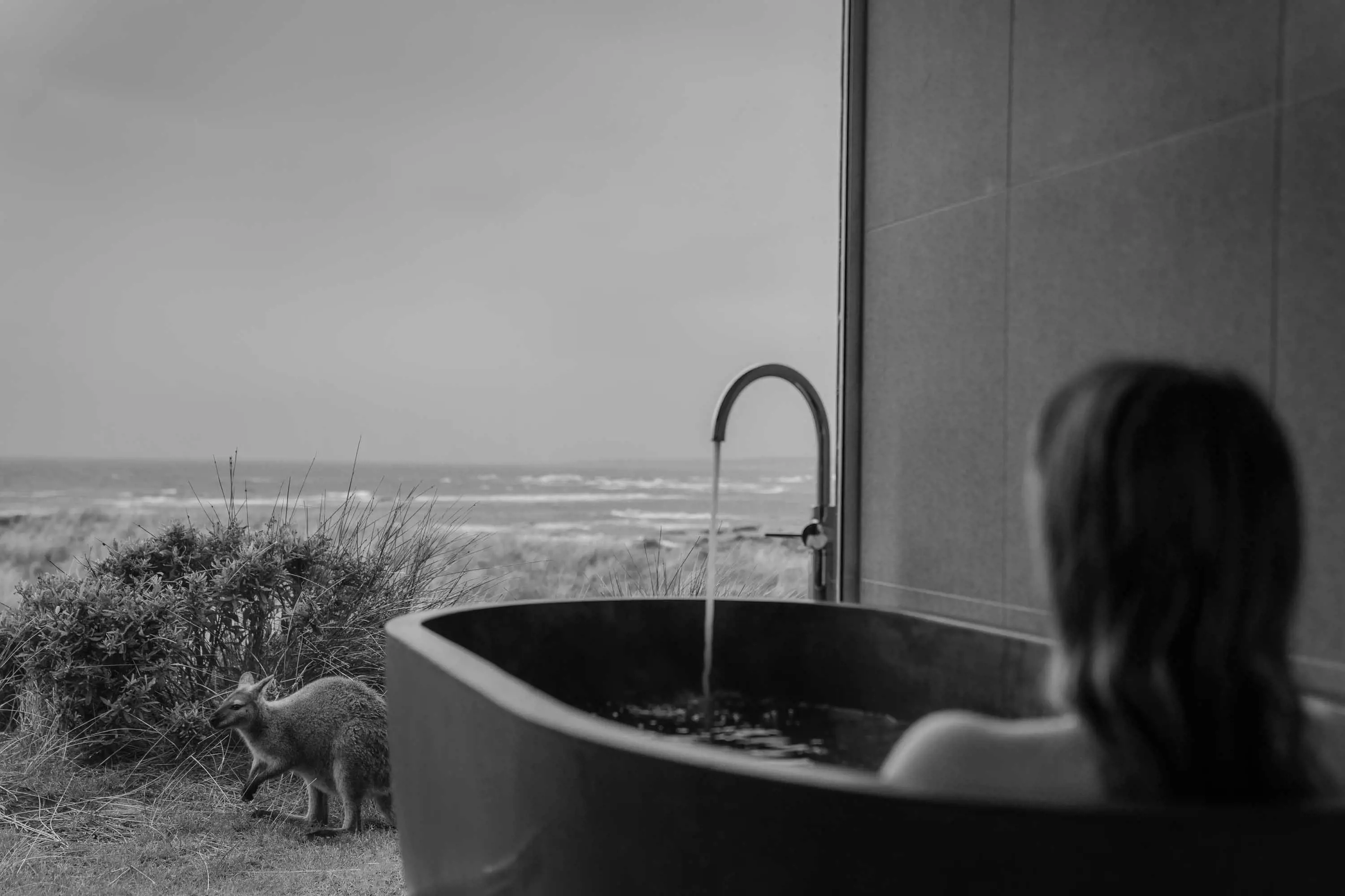 A woman reclines in a large dark stone bath with views of the coast. A small kangaroo grazes in grass nearby.