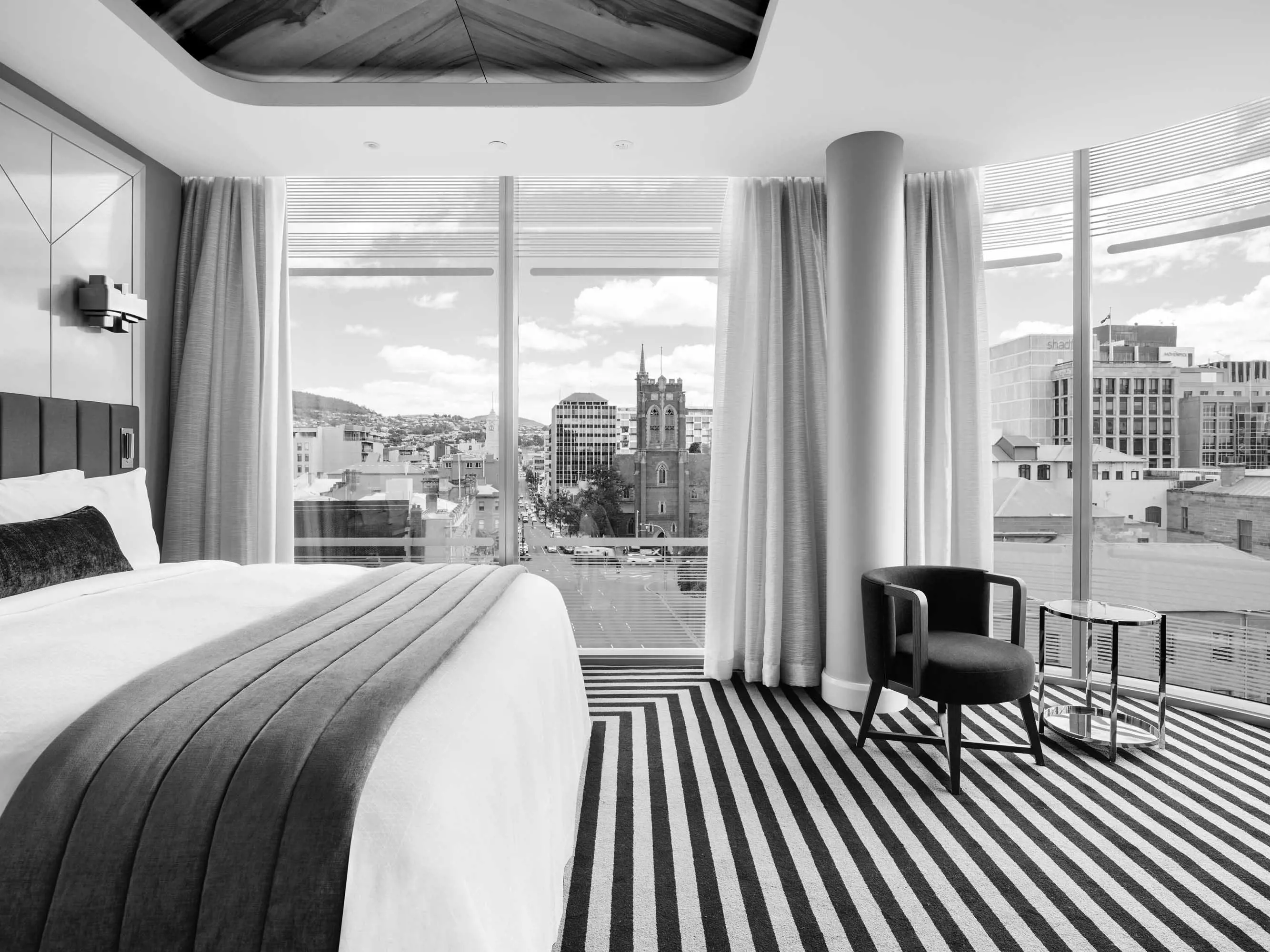 A contemporary hotel room with large windows and elevated views of the Hobart city skyline.
