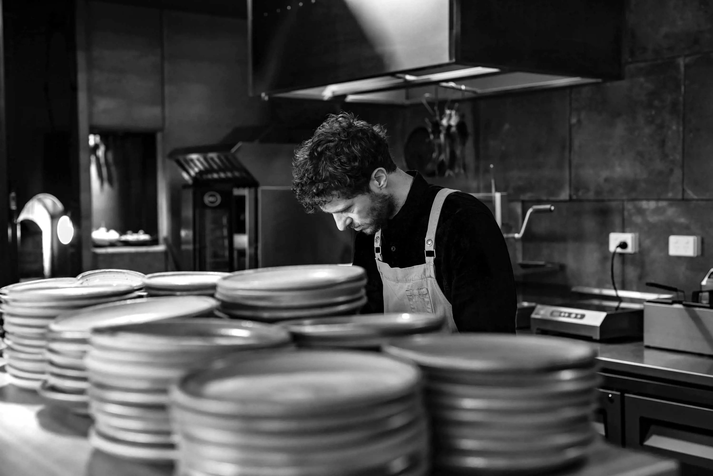 A chef concentrates on preparation behind the counter stacked with plates in a large open kitchen.
