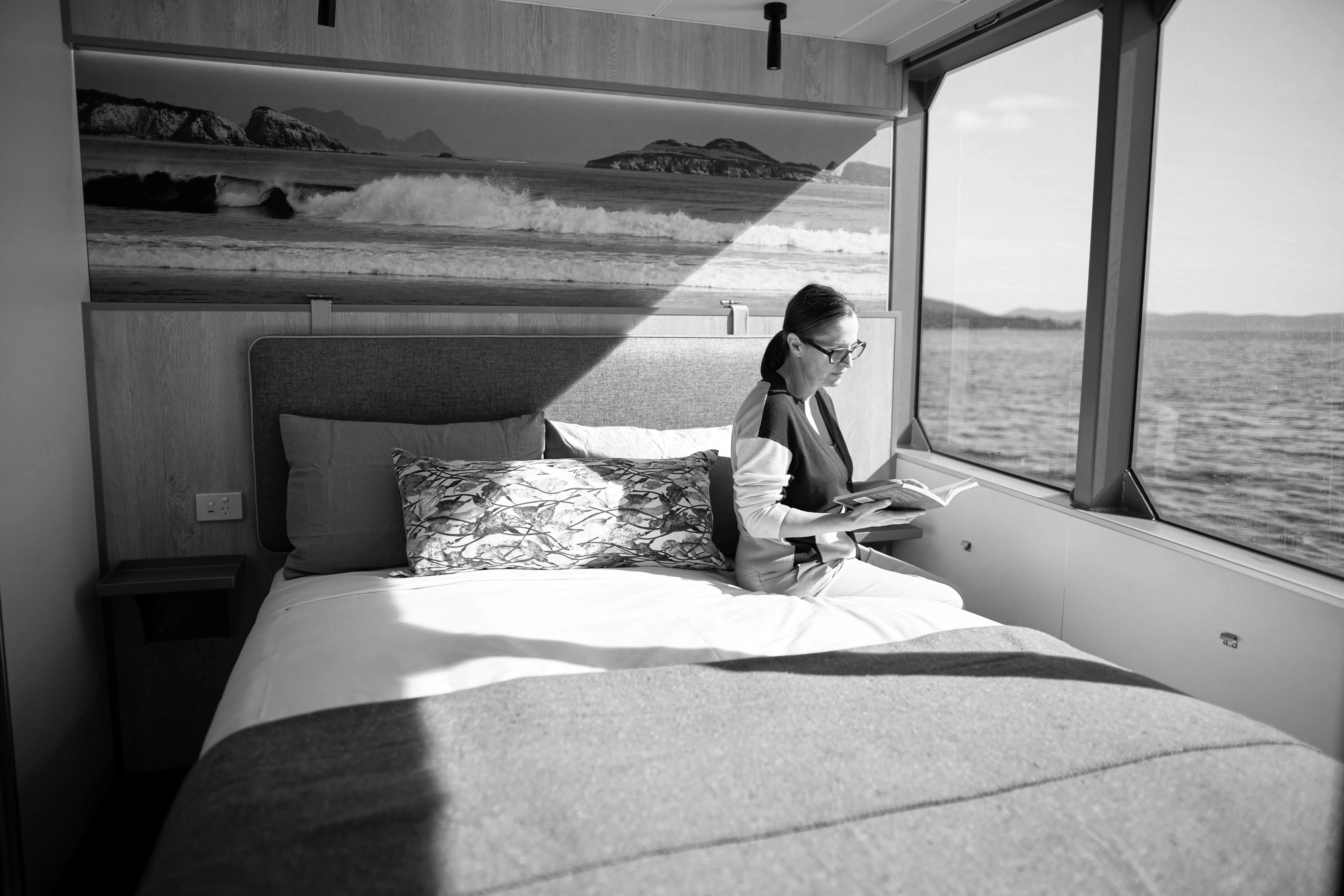 A woman sits on a large bed by the large windows of a boat on calm waters.