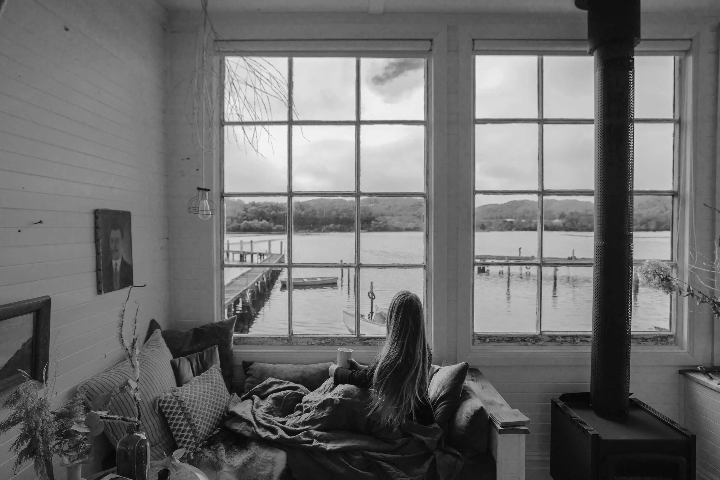 A young woman sits on a couch inside, looking out colonial style windows over water.