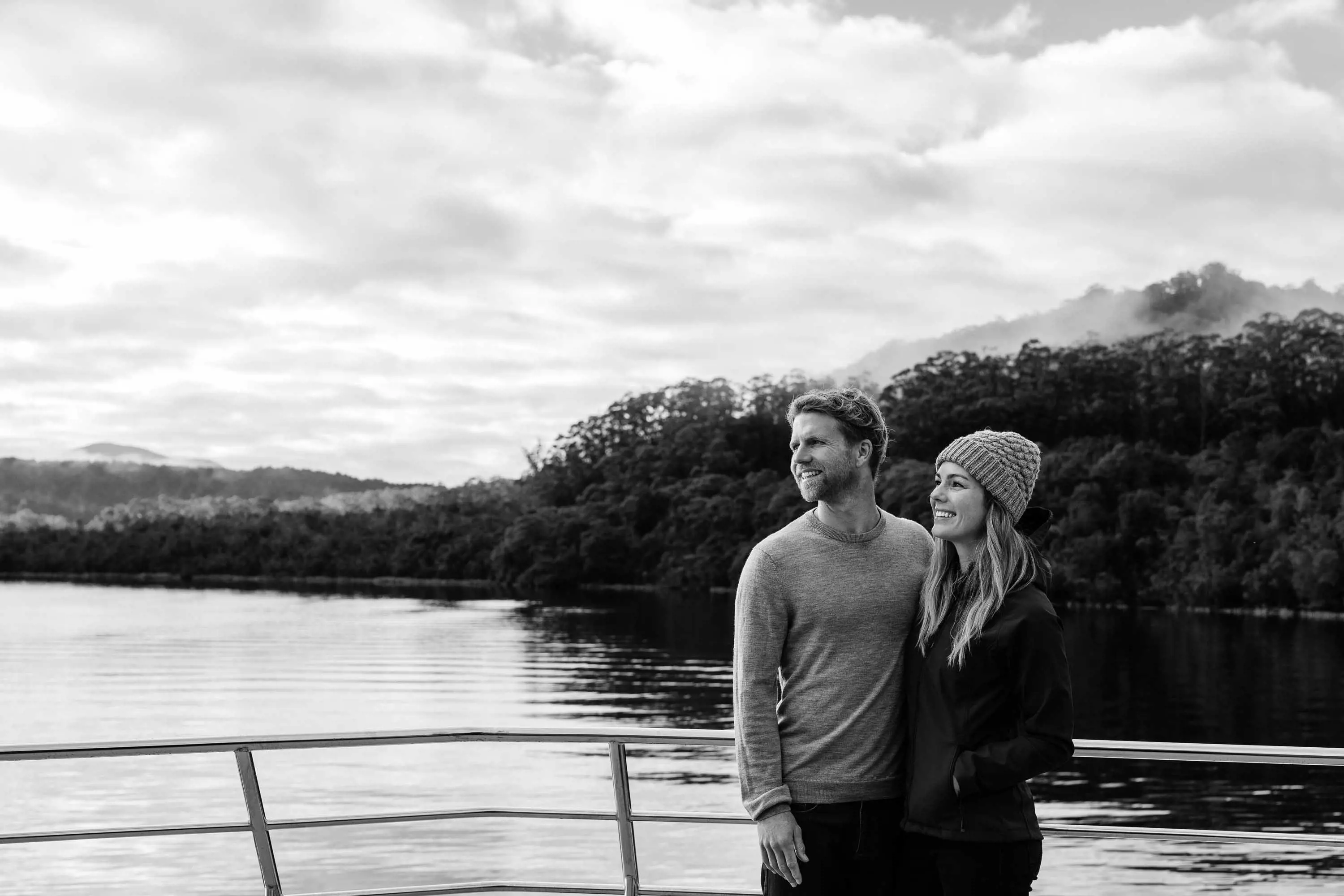 A young couple stand on the bow of a yacht, cruising down the calm waters of a wide river.