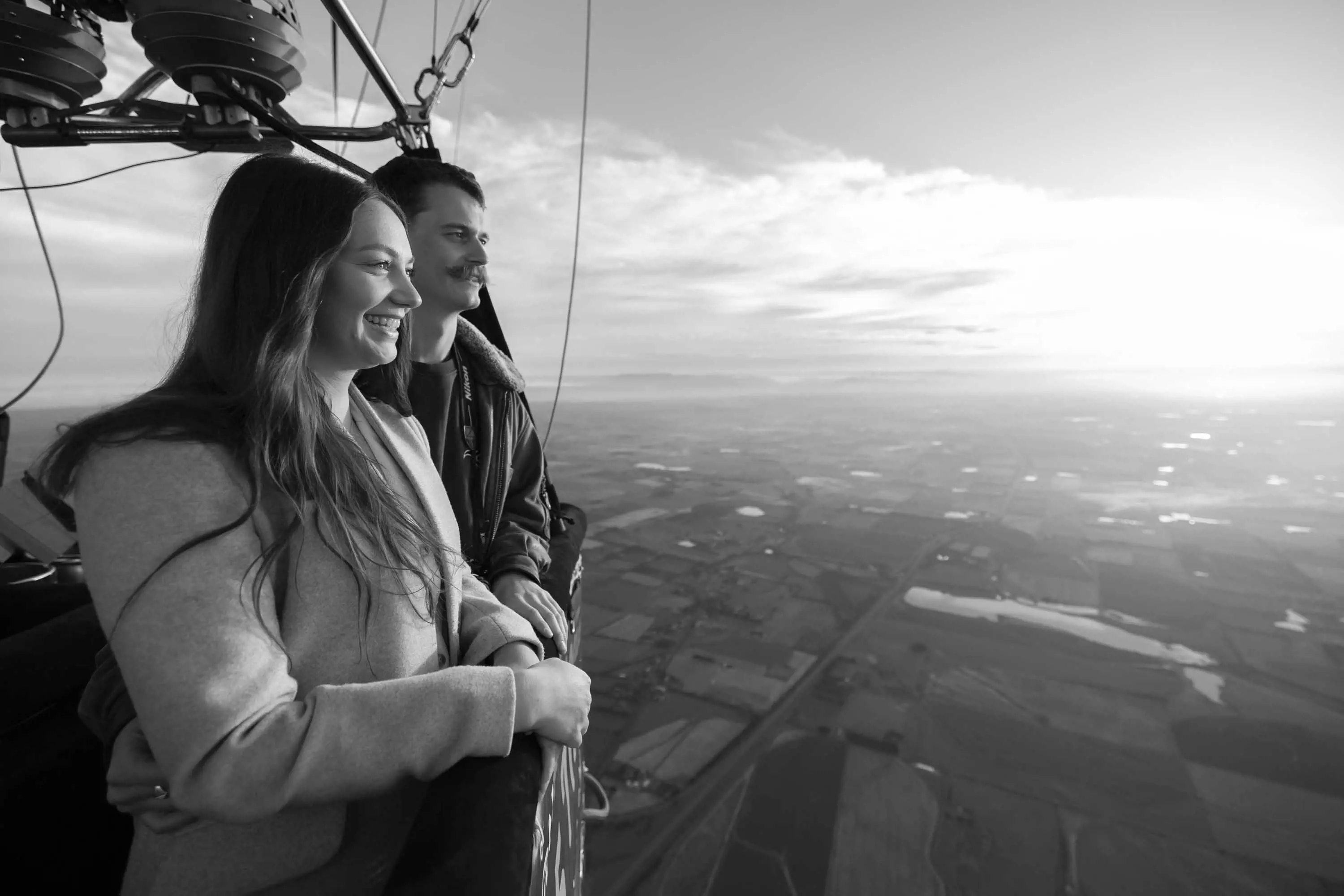 A young couple enjoy the view of farm lands by sunrise from the basket of a large, hot air balloon.