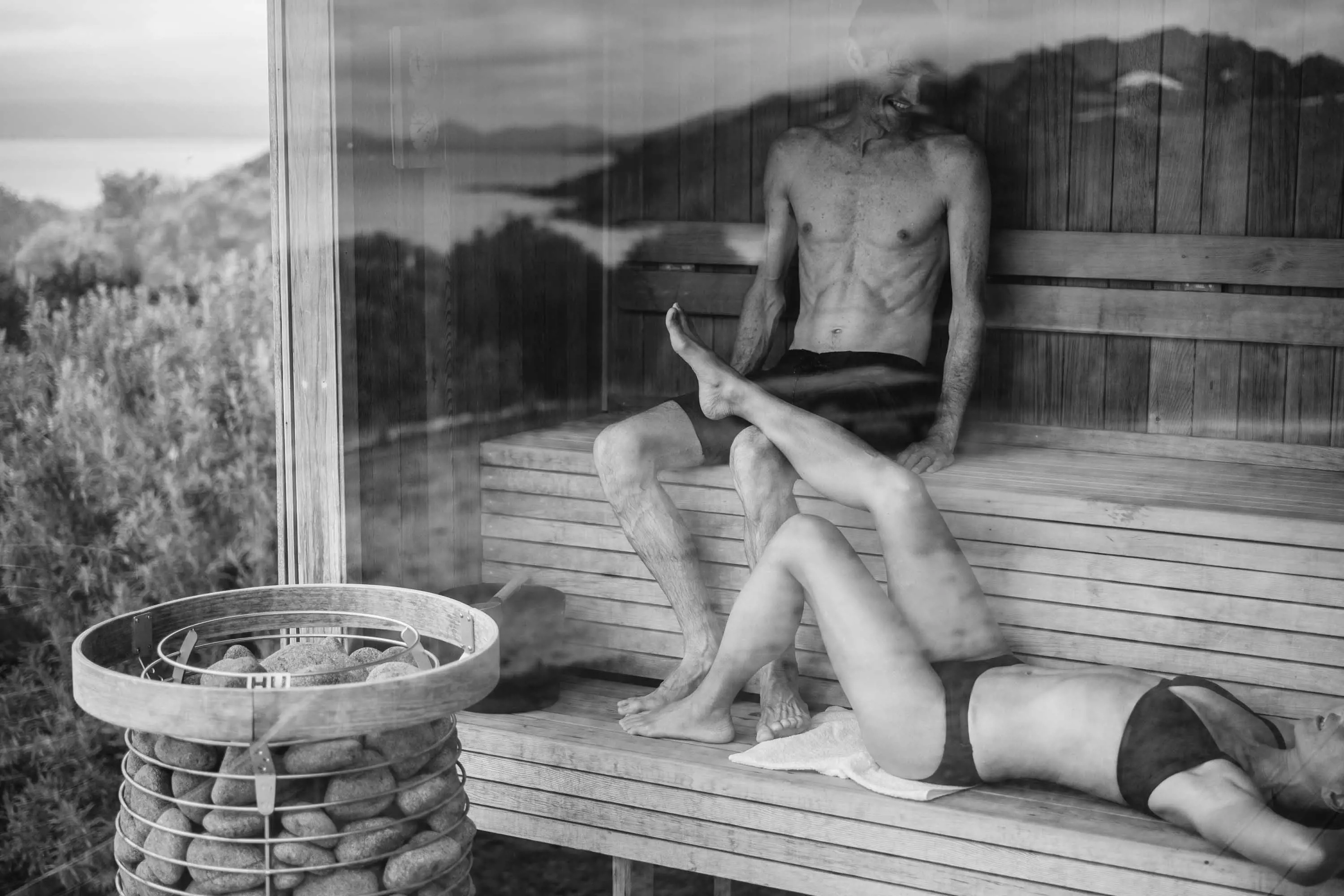 A couple sit inside a sauna with floor-to-ceiling windows overlooking the coast.