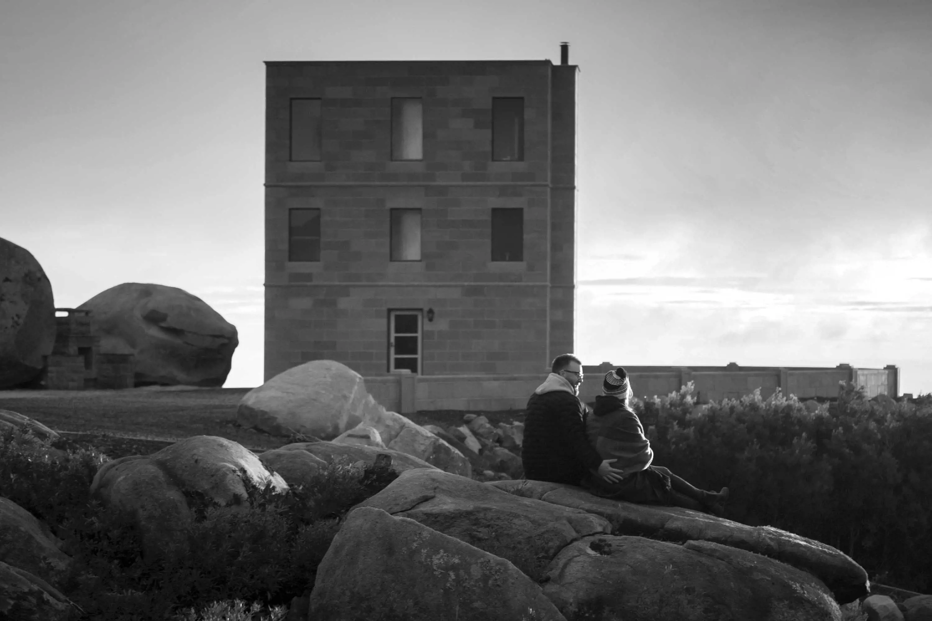 A tall, square, sandstone building stands on a plateau surrounded by large boulders. A young couple are seated in the middle ground on a rock.