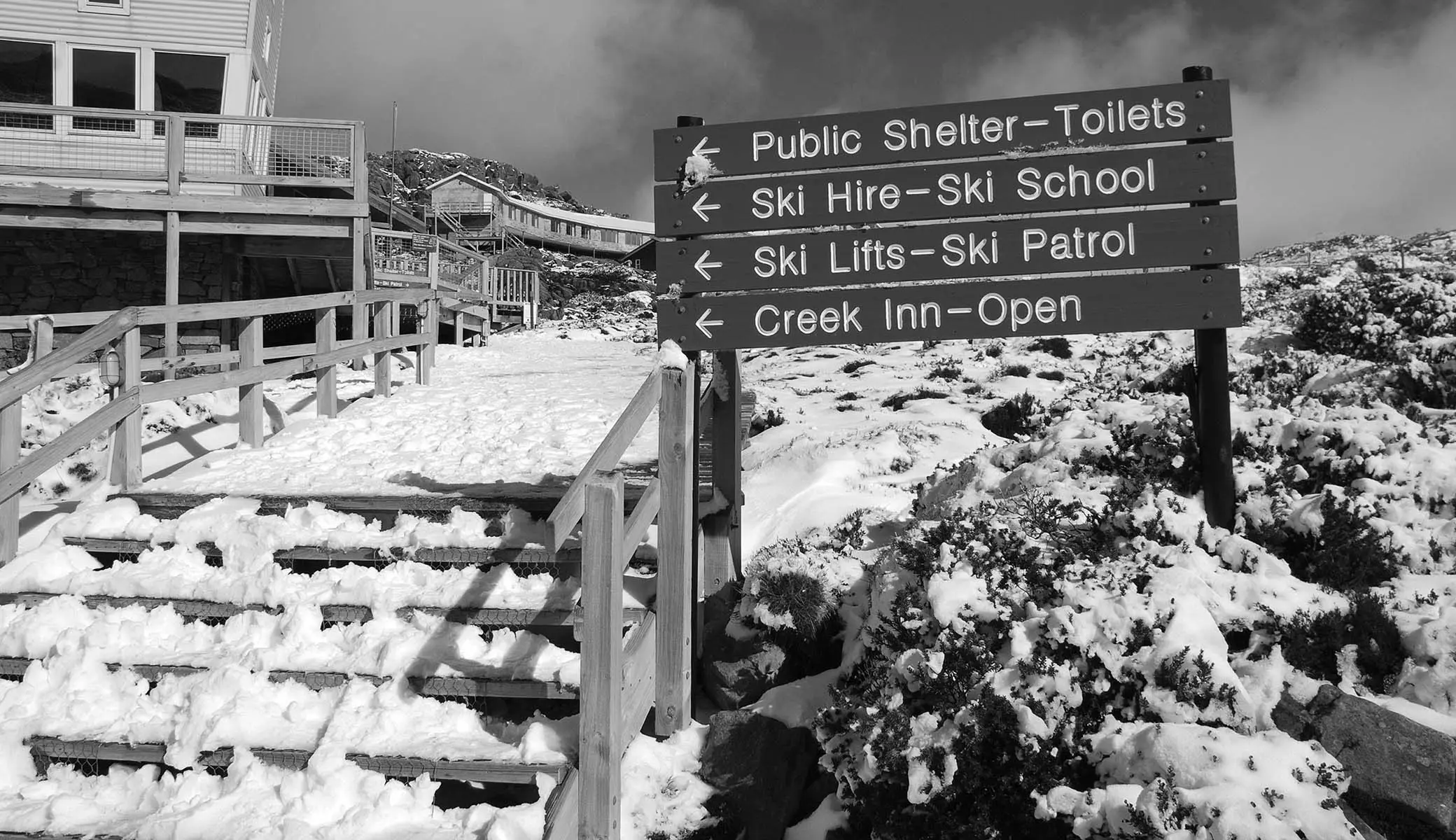 A set of outdoor steps, covered in snow, running alongside chalets. A sign next to the steps points the direction to ski hire, ski school and shelters.