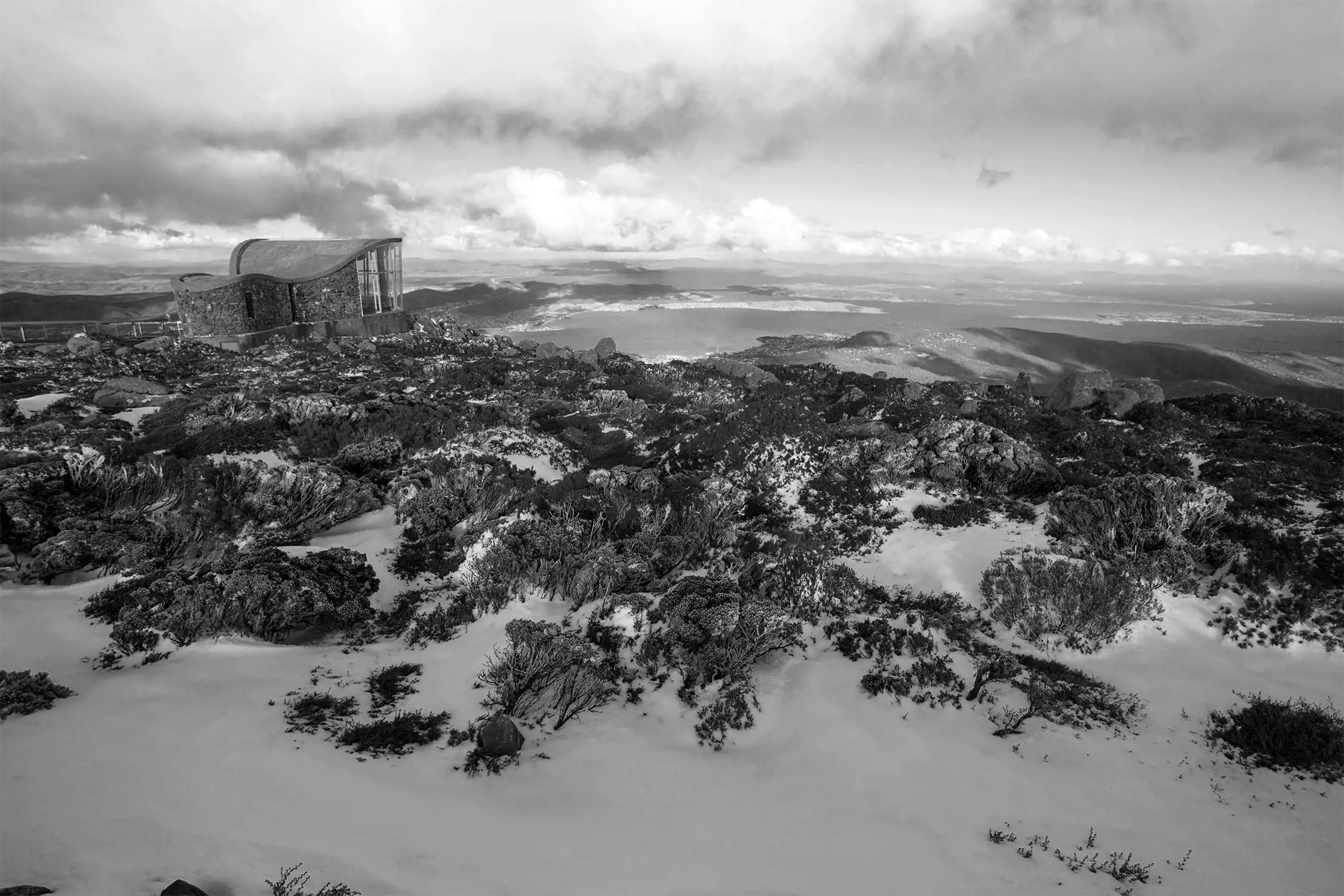A rocky, snow-coverd landscape showcases a scenic building at a high elevation under a cloudy sky.