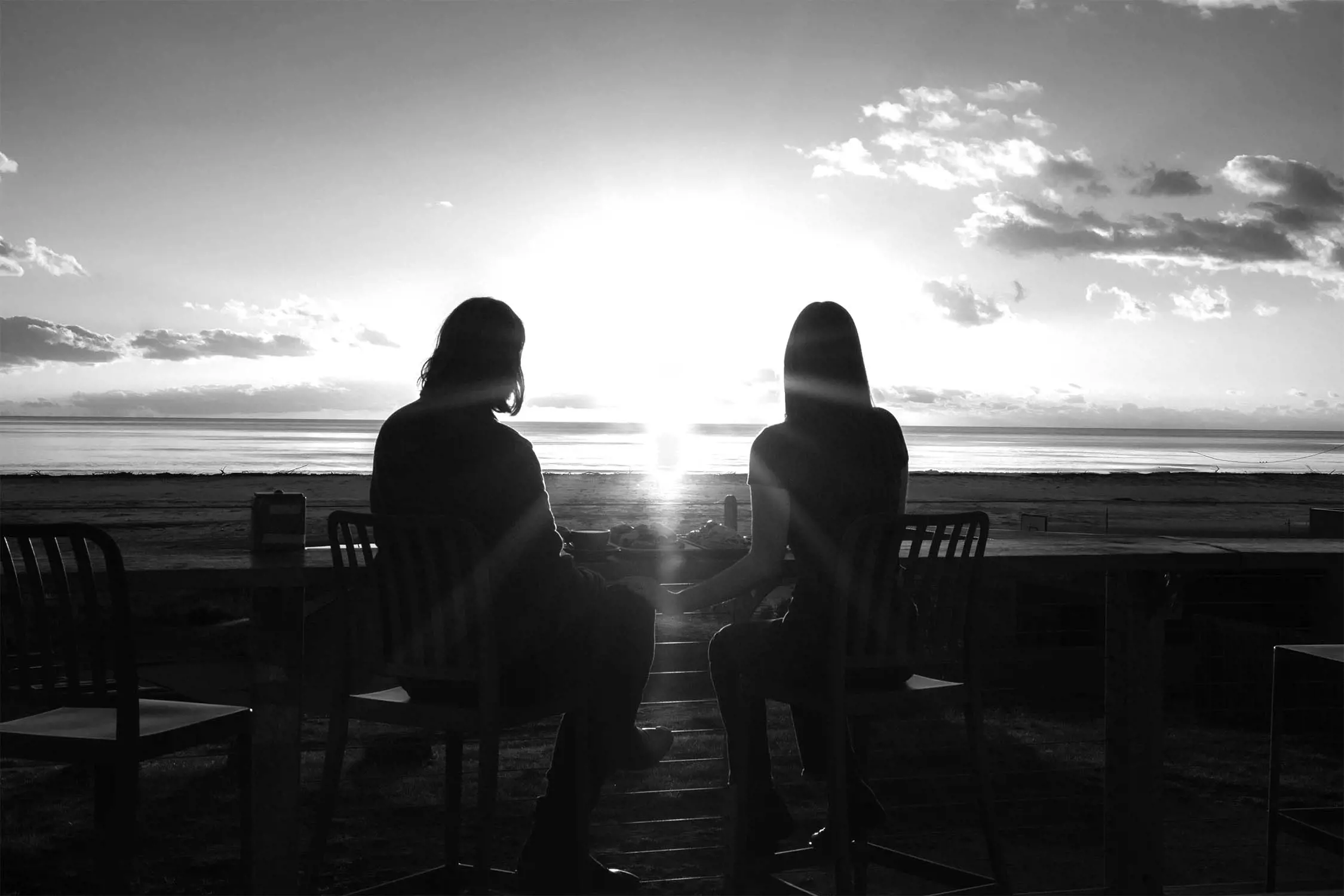Two silhouetted figures sit together at a table, watching the sunset over the water.