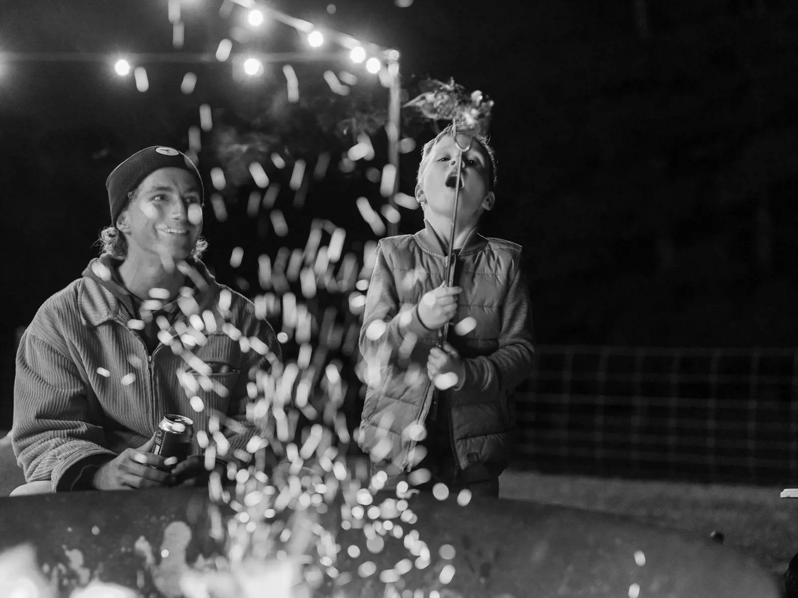 A man and child sit over a large firepit, which is sending up sparks. The boy blows out the fire on the end of a roasting stick.