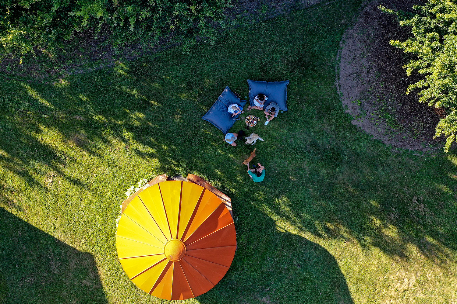 A top-down view of picnic makers sitting under an umbrella on a large grassy area.