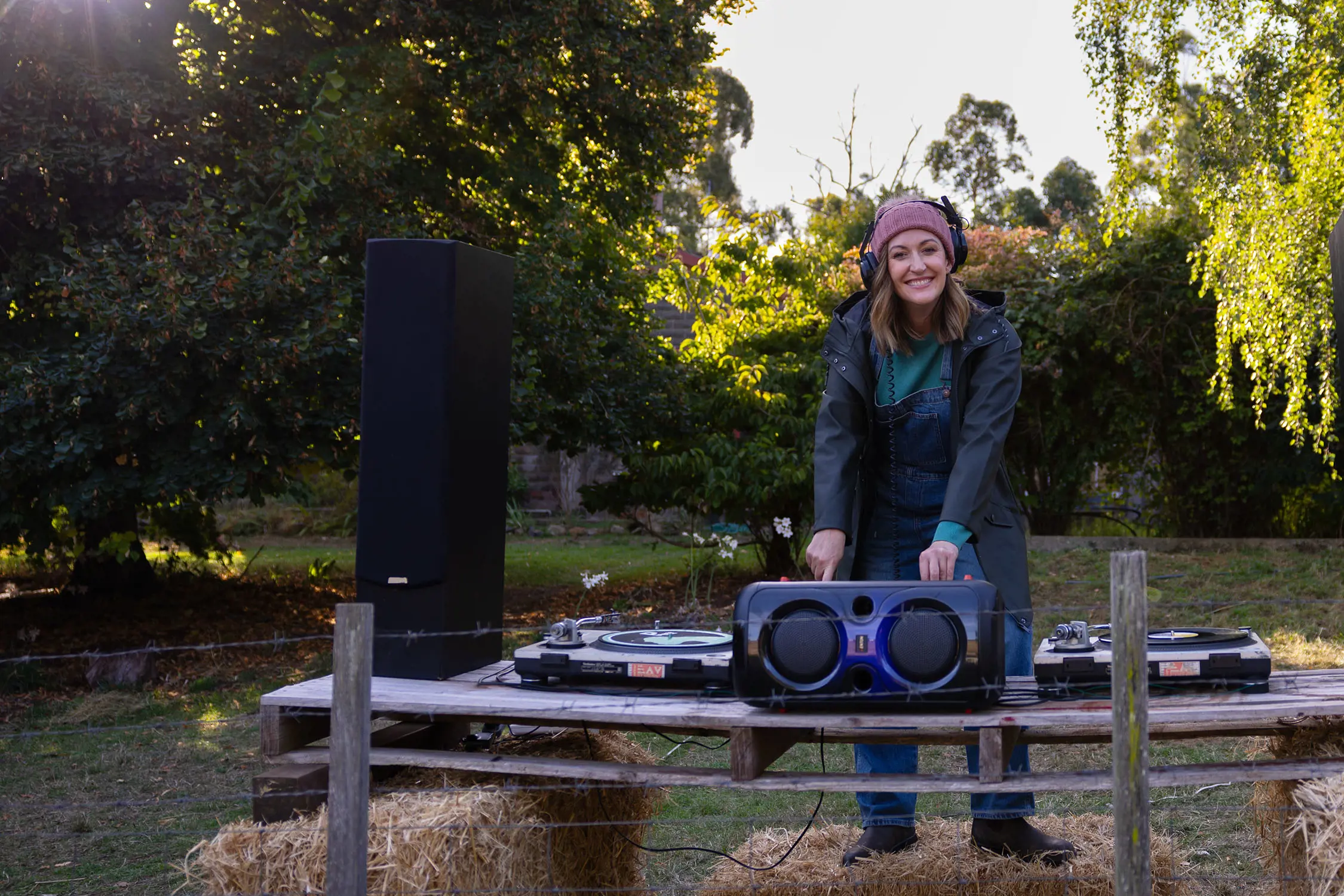 A smiling woman wearing headphones and a rain jacket, standing behind a DJ desk with turntables and speakers set up on bales of hay outdoors.