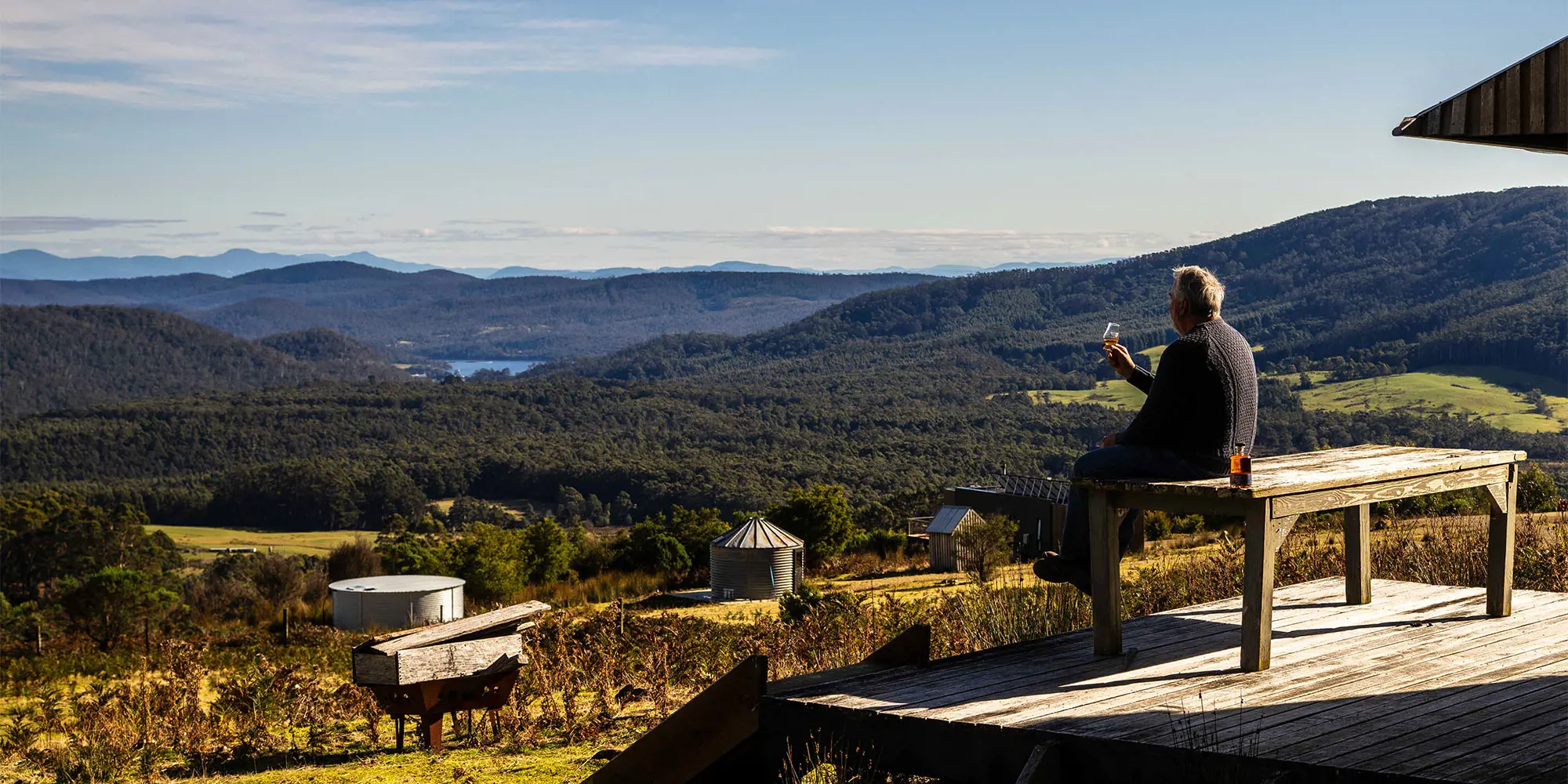 Person sitting on a wooden deck beside a rustic cabin, holding a wine glass while overlooking a valley filled with vineyards, forests and hills.