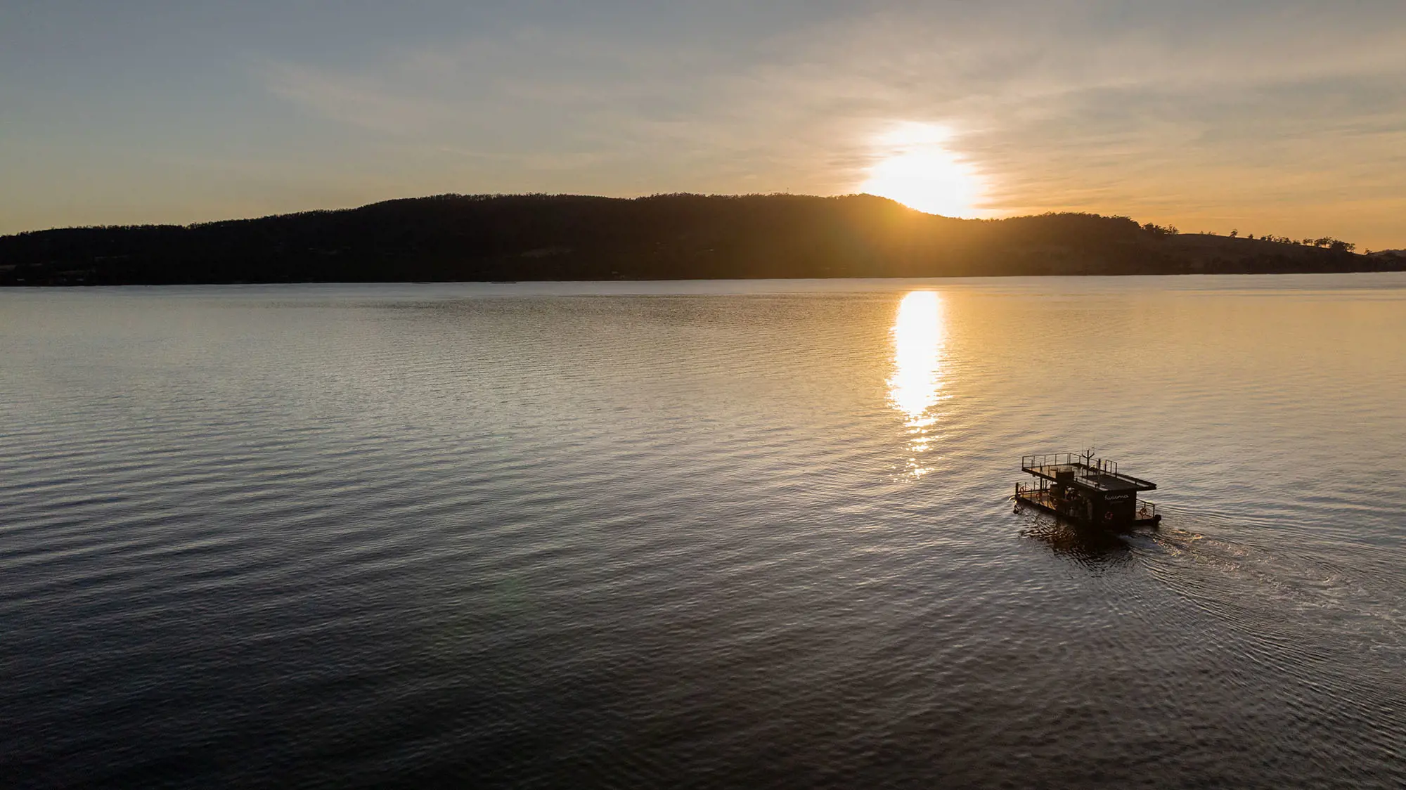 A floating sauna drifting on a calm lake at sunset with hills and a clear sky in the background.