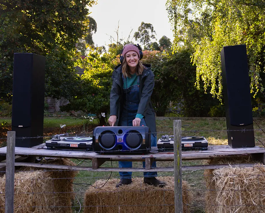 A smiling woman wearing headphones and a rain jacket, standing behind a DJ desk with turntables and speakers set up on bales of hay outdoors.