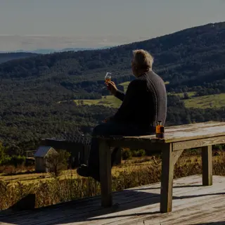 Person sitting on a wooden deck beside a rustic cabin, holding a wine glass while overlooking a valley filled with vineyards, forests and hills.