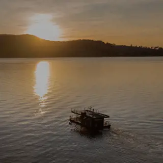 A floating sauna drifting on a calm lake at sunset with hills and a clear sky in the background.