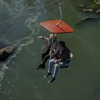 Two people seated on a red chairlift, suspended crossing above a rocky river surrounded by trees.