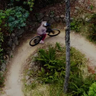 An aerial view of a mountain biker, riding on a narrow winding dirt trail through dense forest.