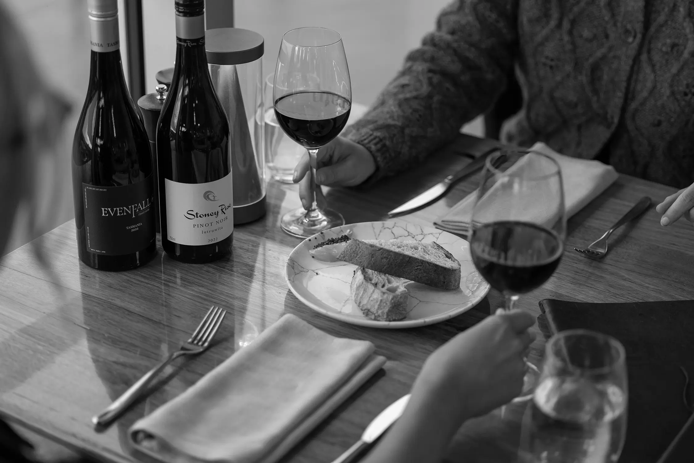 Looking over the shoulders of two people seated at a restaurant table. A shared plate of bread is in front of them and red wine fills the wine glasses.